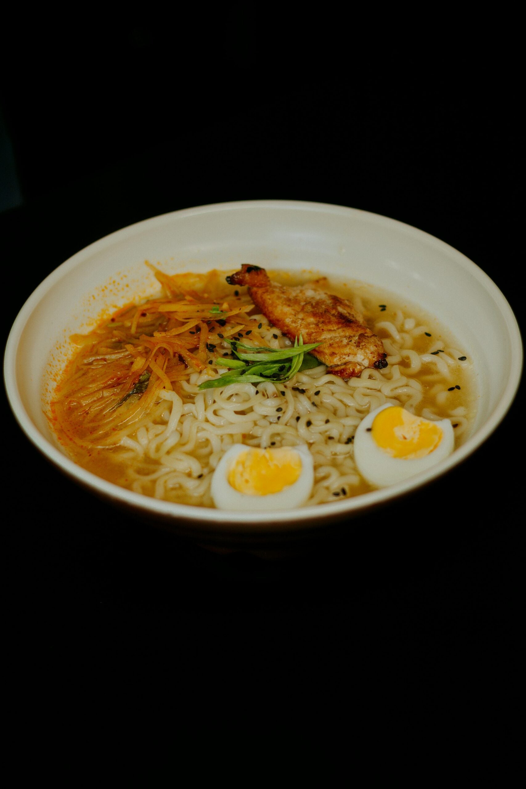 A bowl of ramen with sliced grilled chicken, shredded carrots, green onions, and two halved boiled eggs, served in a light broth against a dark background.