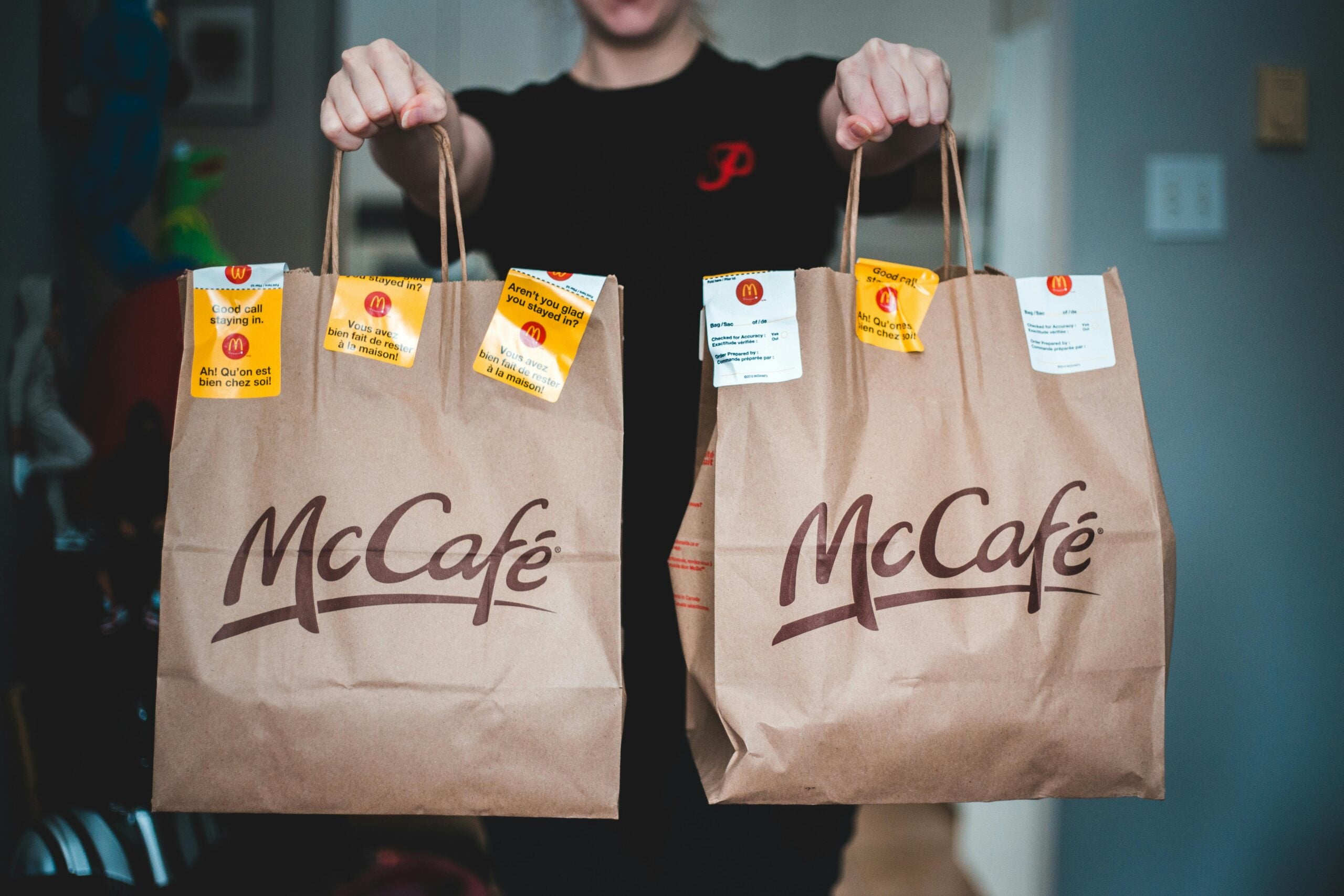 A person holds up two McCafé paper bags with order stickers on them, standing indoors. The background is slightly blurred, focusing on the bags and the person's hands.