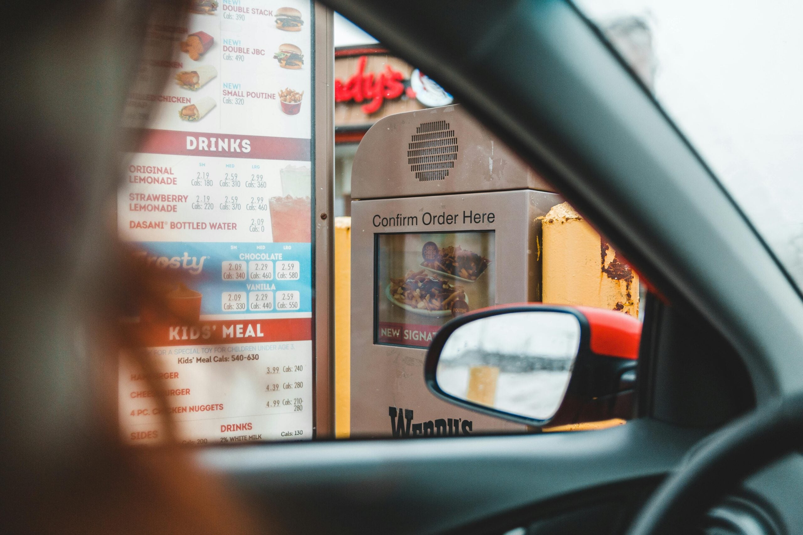 A drive-thru menu and order speaker at a fast-food restaurant are seen through a car window and side mirror. The display reads “Confirm Order Here.” The restaurant branding and menu items are partially visible.