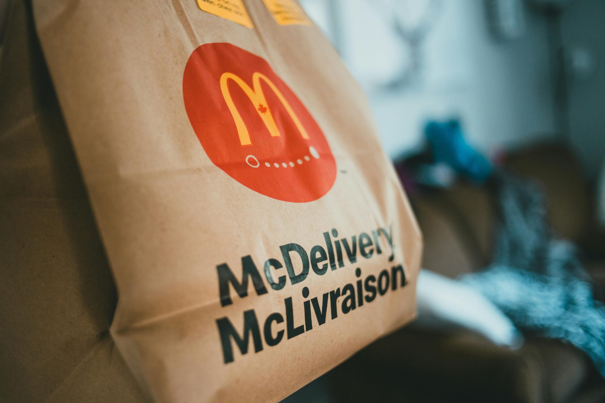 A close-up of a brown paper McDonald's delivery bag with "McDelivery" and "McLivraison" printed on it, featuring the McDonald's logo. The background is blurred, showing an indoor setting.