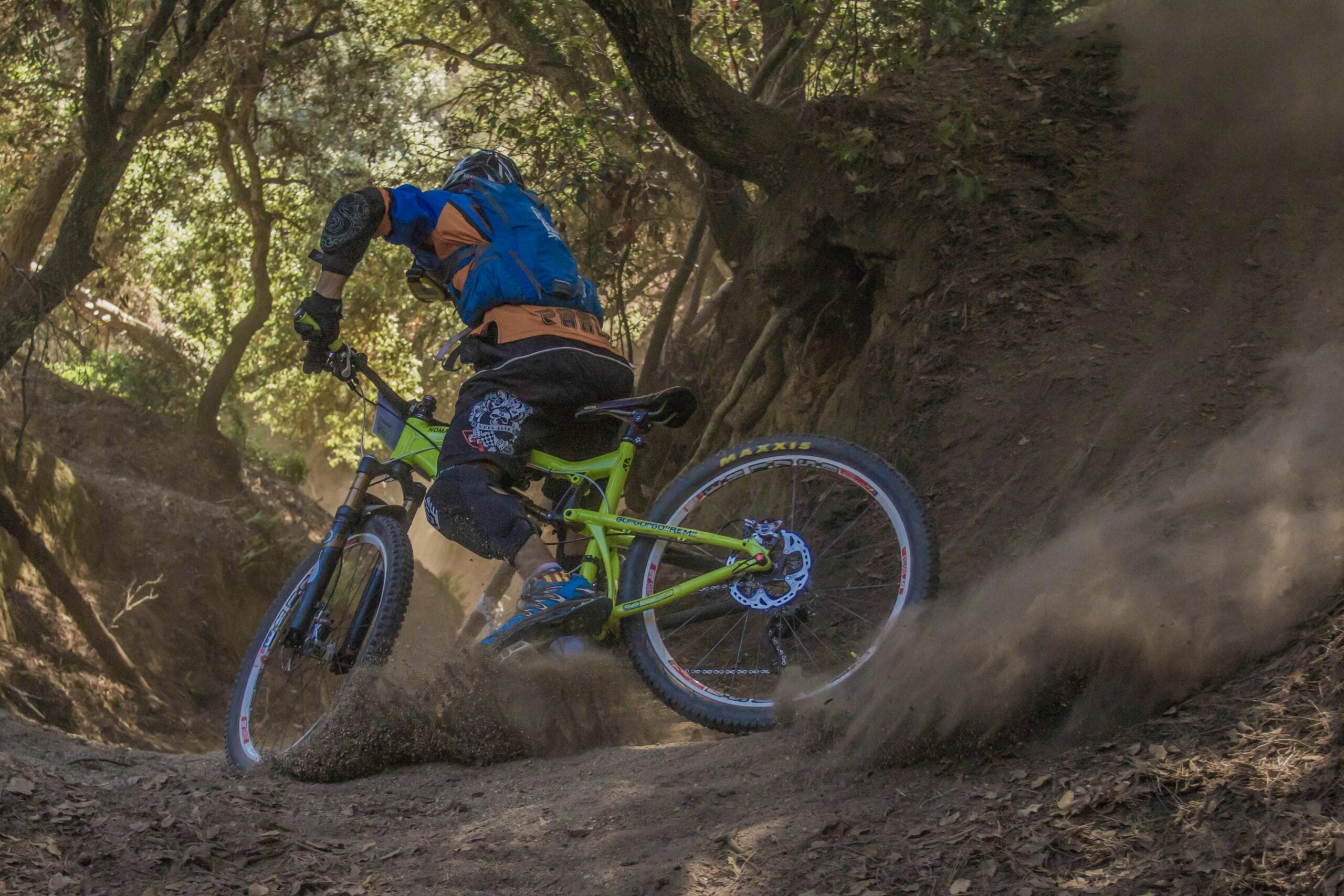 A mountain biker wearing protective gear rides downhill on a dirt trail through a forest, kicking up a cloud of dust as they navigate a sharp turn.