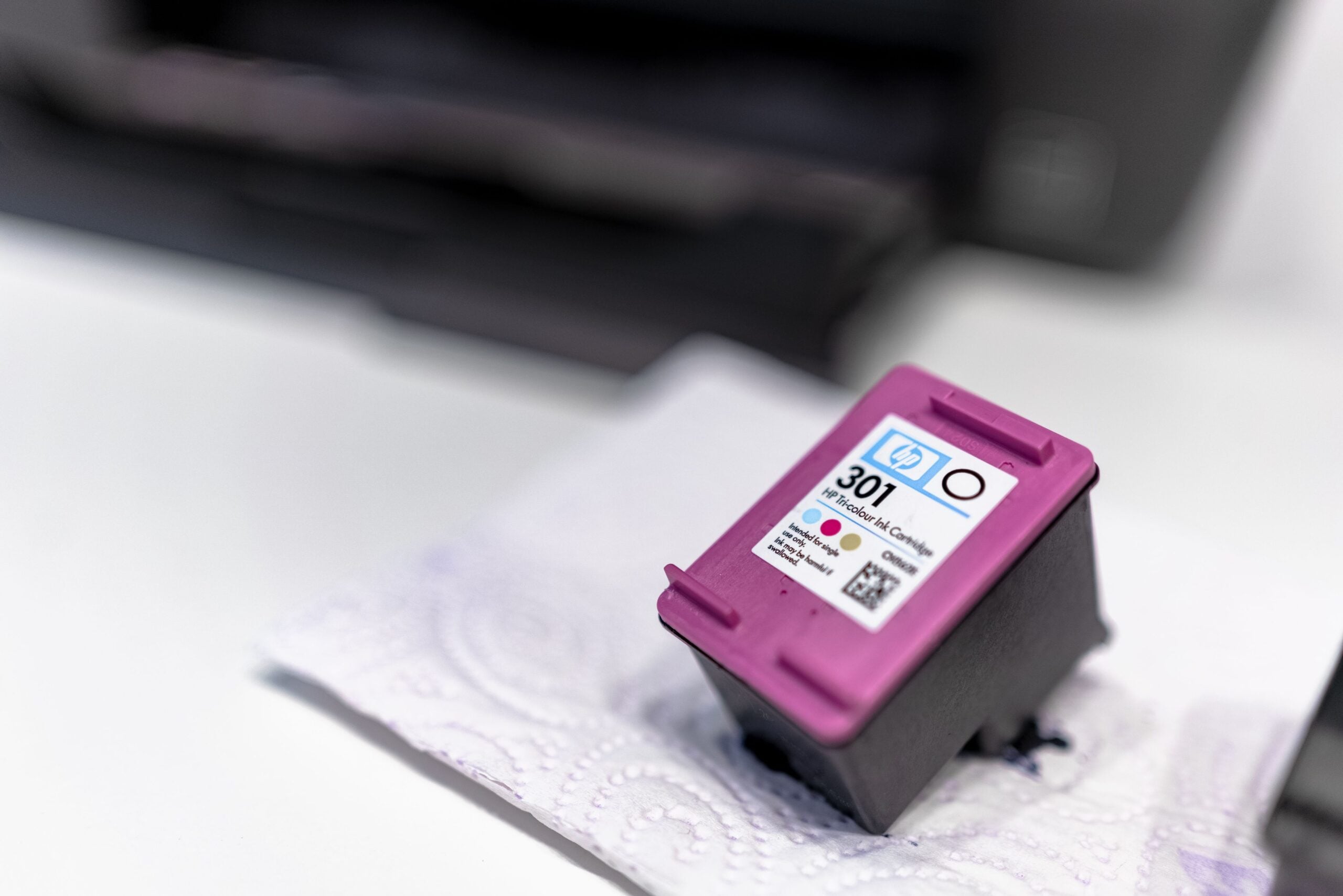 A close-up of a tri-color HP 301 ink cartridge resting on a paper towel with a blurred printer in the background. The cartridge has a magenta top and visible labels indicating color and brand.