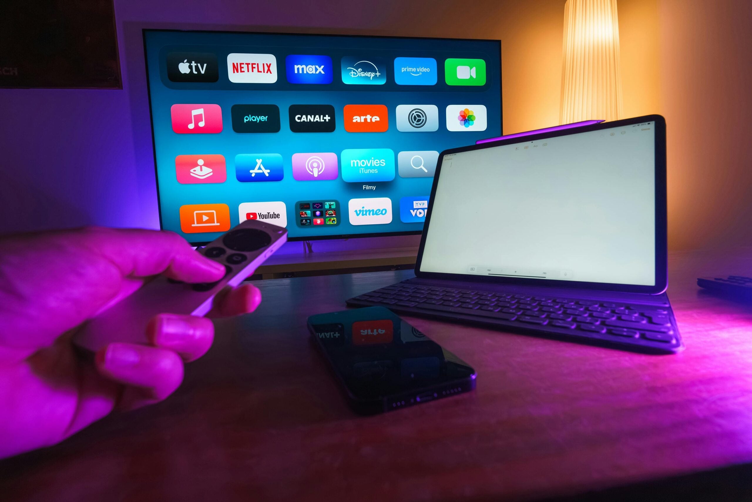 A person holds a remote in front of a TV displaying various streaming apps. A tablet with a keyboard and a smartphone sit on the desk nearby, all illuminated by colorful ambient lighting.