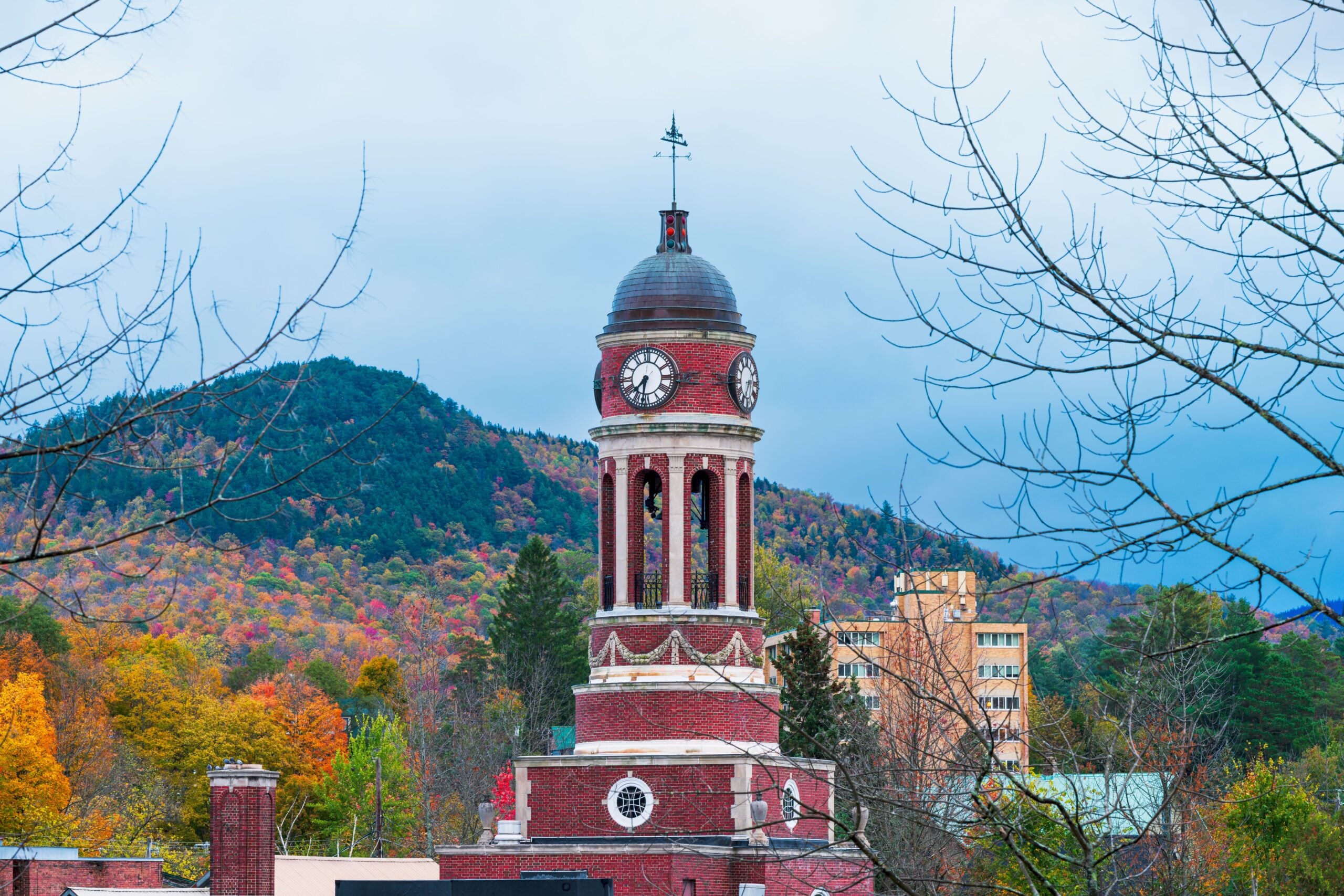 A red-brick clock tower with a dome stands among autumn trees, with a forested mountain in the background under a cloudy sky.