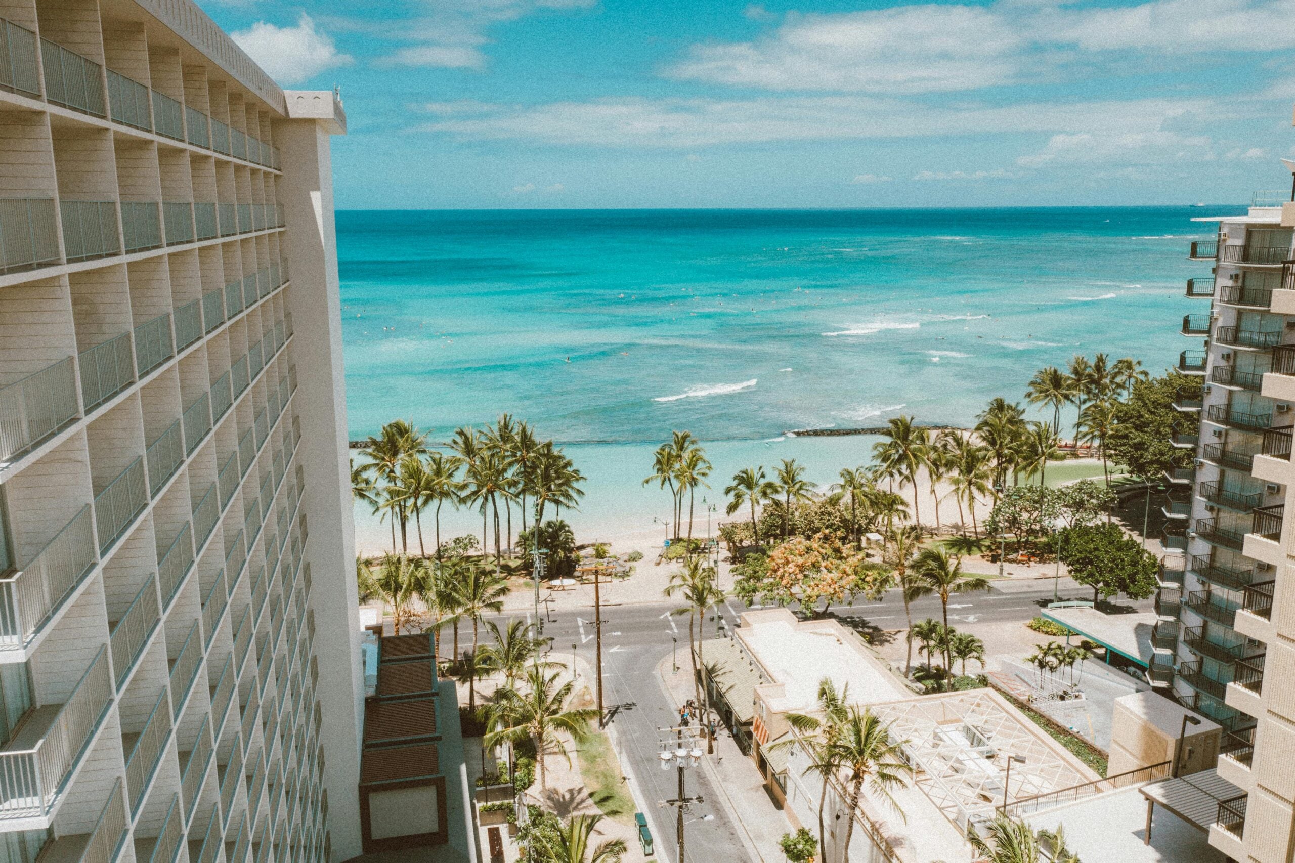 A bright, sunny view of a tropical beach with turquoise water, palm trees, and a sandy shore, seen from between two tall hotels with balconies. A road and lush greenery are visible below.