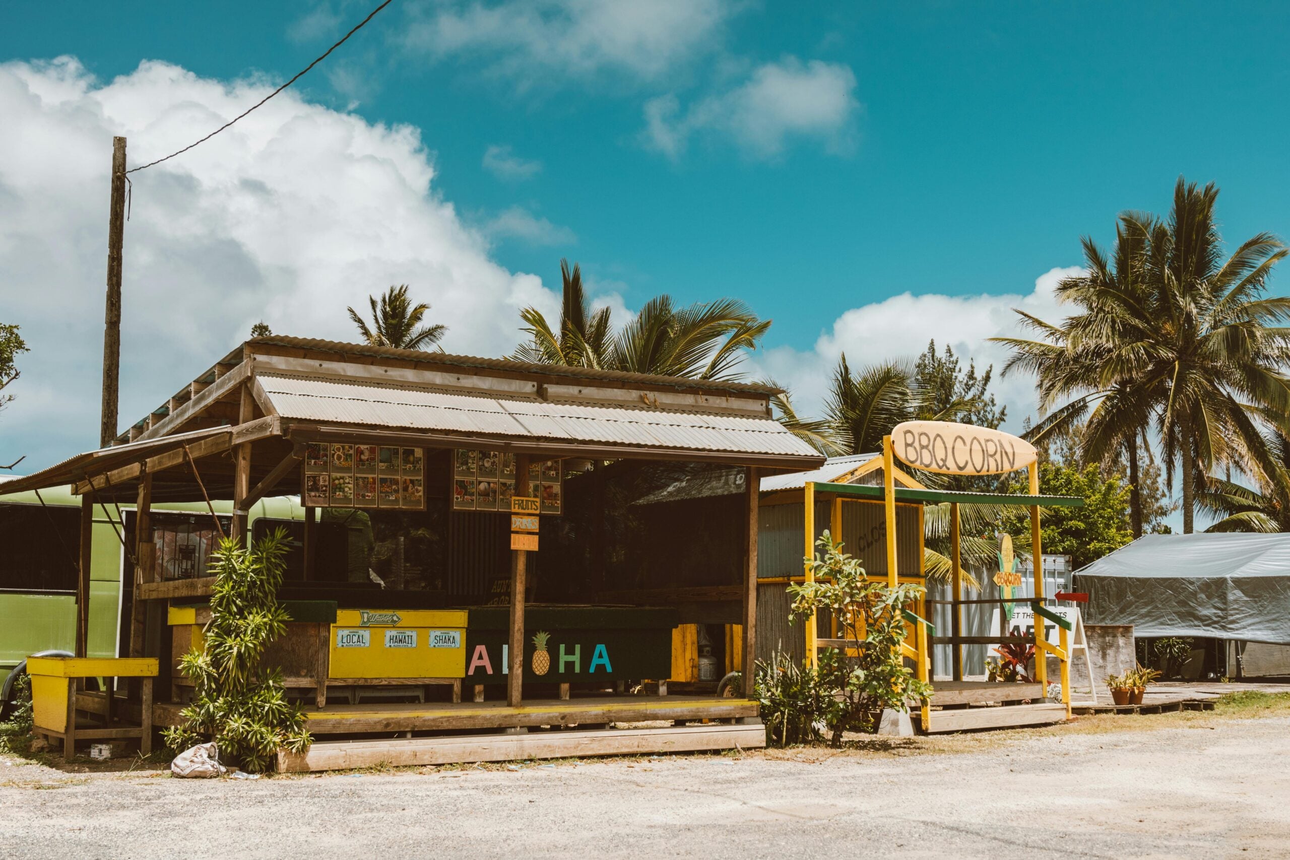 A small outdoor food stand with a wooden roof and “Aloha” sign stands next to a yellow BBQ Corn shack, surrounded by palm trees under a bright blue sky with fluffy clouds.