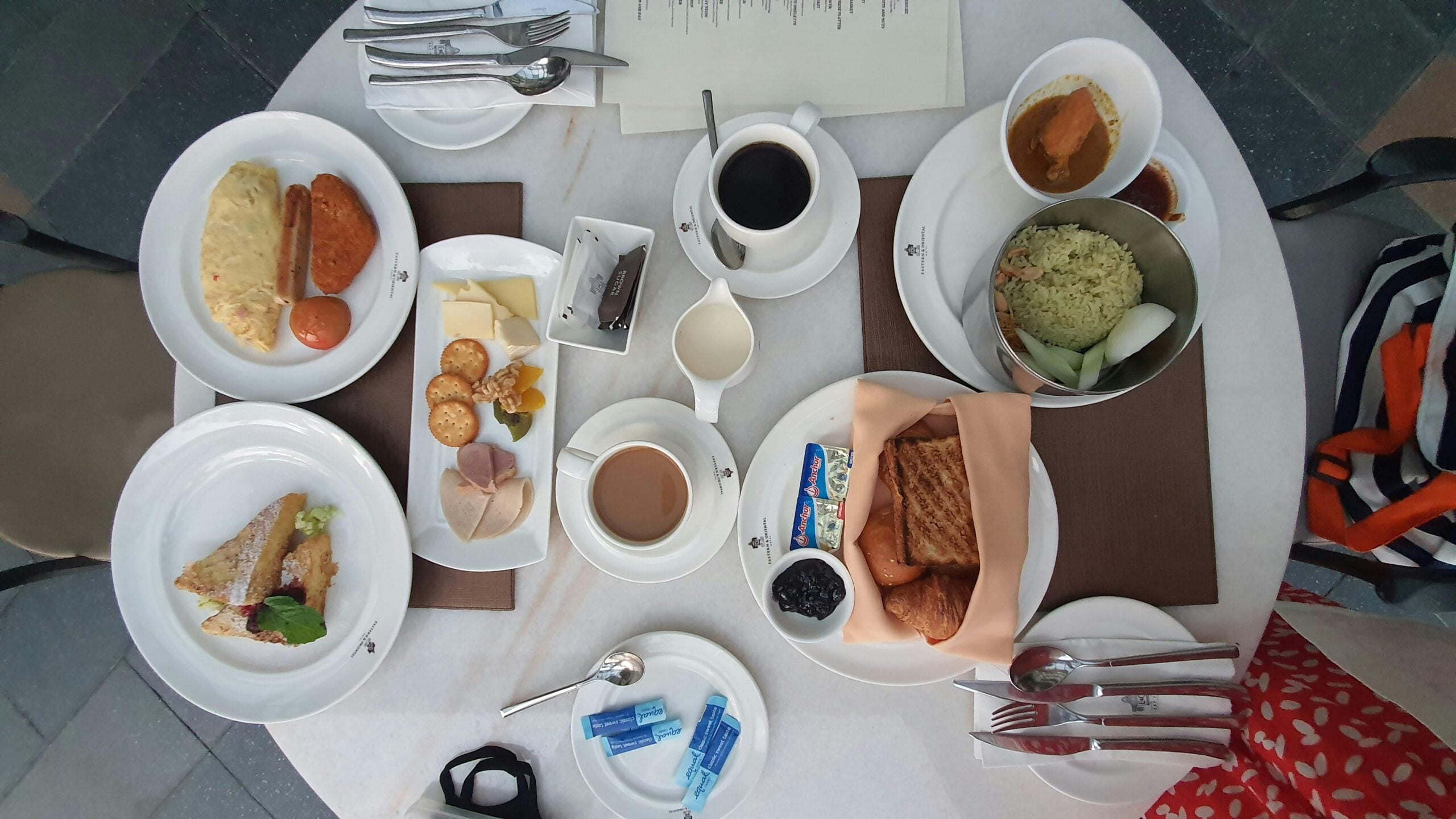 A breakfast table set for two with plates of toast, omelet, sausages, sandwiches, cheese, crackers, fruit, tea, coffee, and condiments. Cutlery and napkins are neatly arranged on brown placemats.