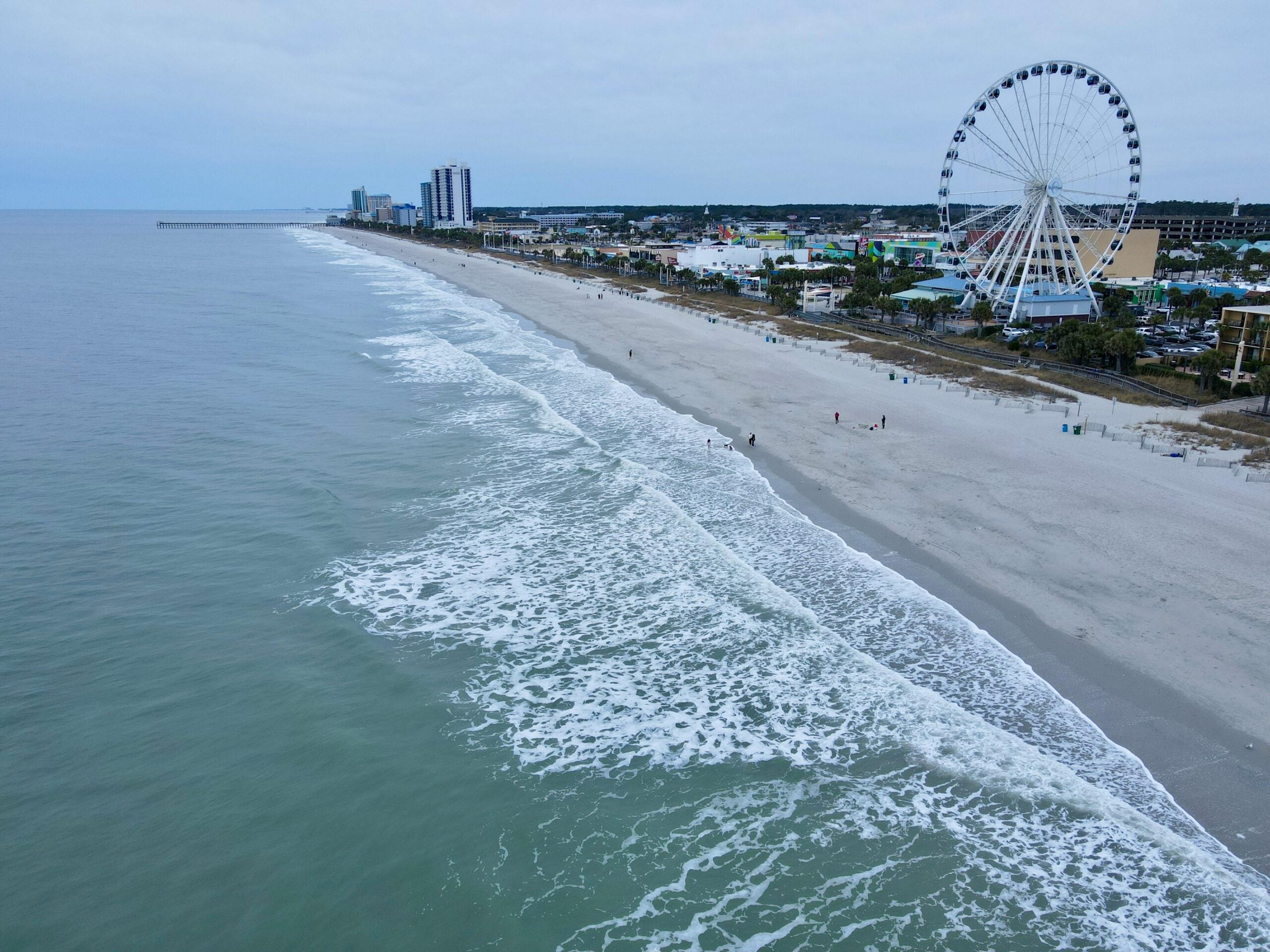 A wide sandy beach with gentle ocean waves, a few people walking along the shore, and a large Ferris wheel and tall buildings in the background under a cloudy sky.