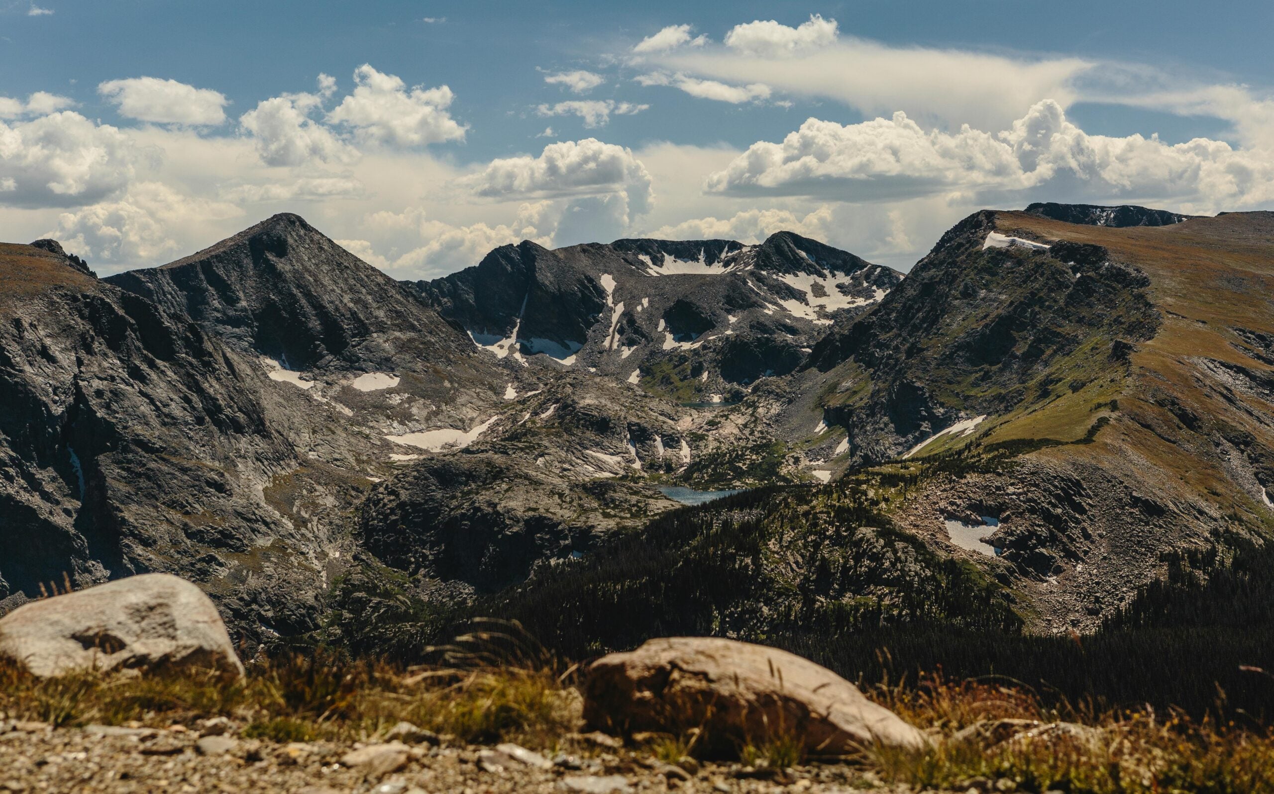 Rocky mountain landscape under a partly cloudy sky, with rugged peaks, patches of snow, rocky terrain, and sparse vegetation in the foreground.