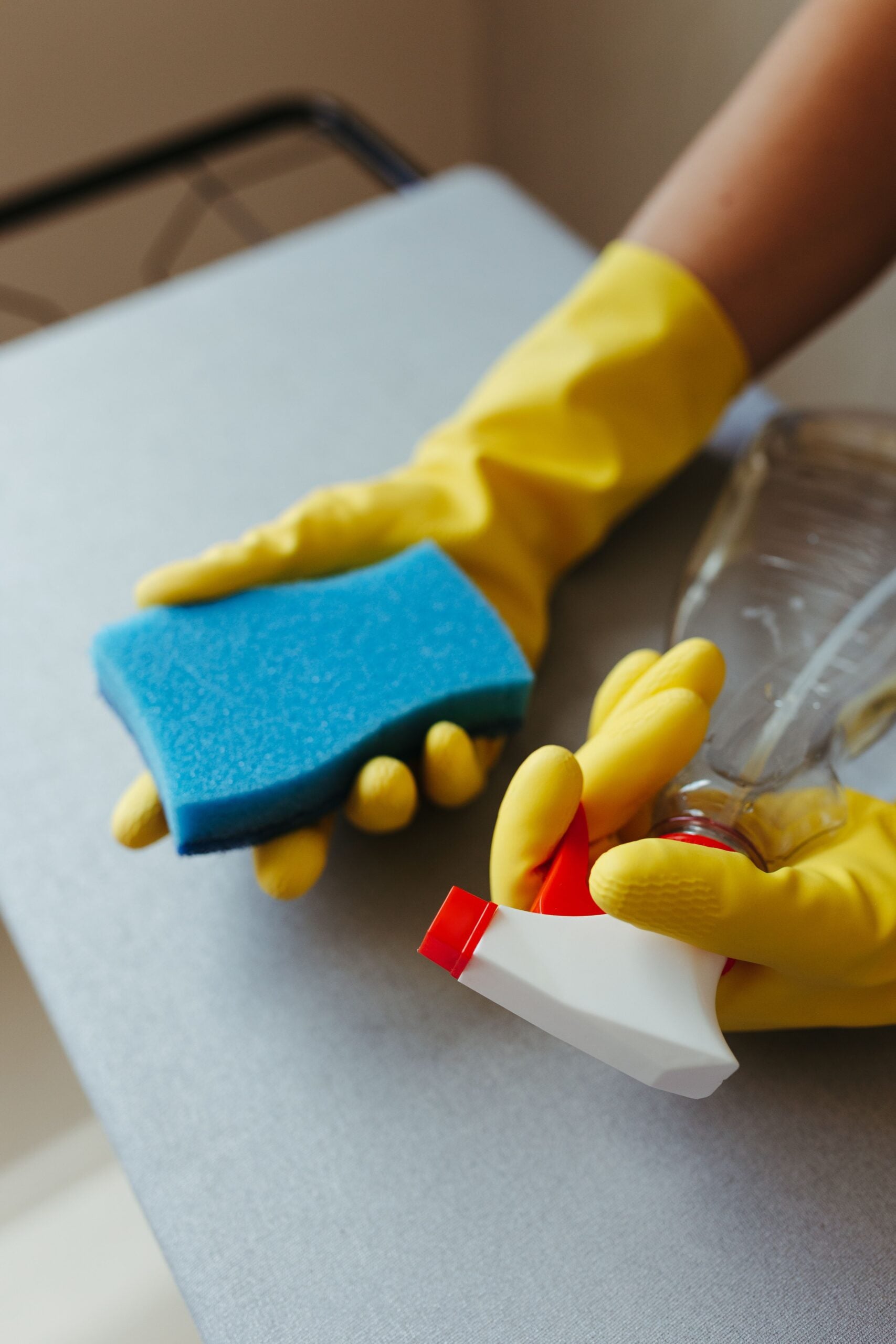 A person wearing yellow rubber gloves holds a blue sponge in one hand and a clear spray bottle with a red nozzle in the other, cleaning a grey surface.