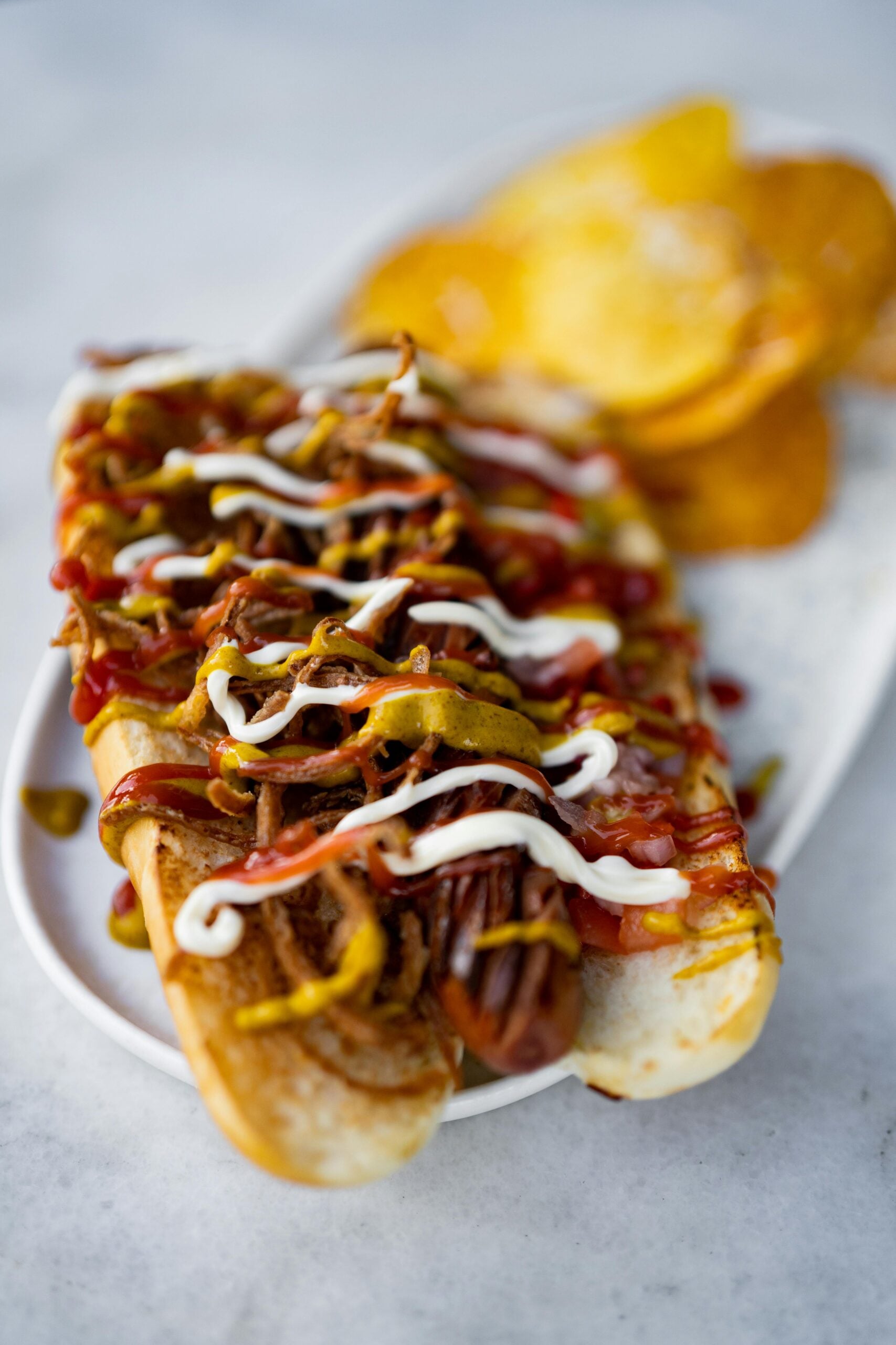 A close-up of a loaded hot dog topped with pulled meat, ketchup, mustard, and mayonnaise on a white plate with crispy potato chips in the background.