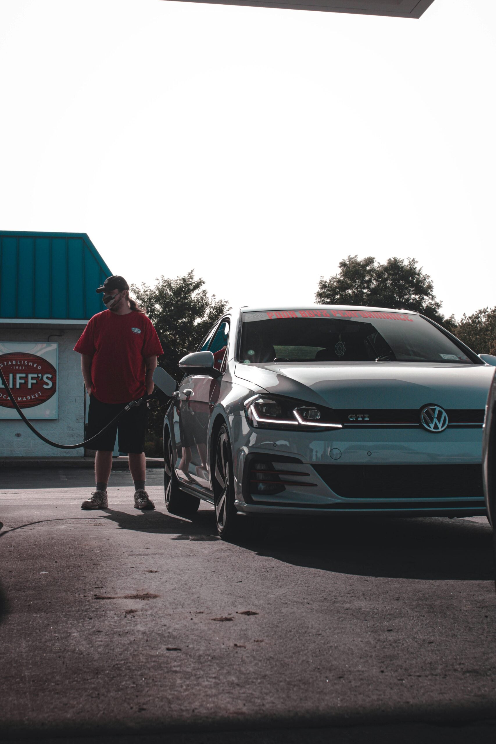 A person wearing a red shirt and black shorts stands near a silver Volkswagen GTI at a gas station, holding a fuel pump. The background shows a building with a sign and some trees.