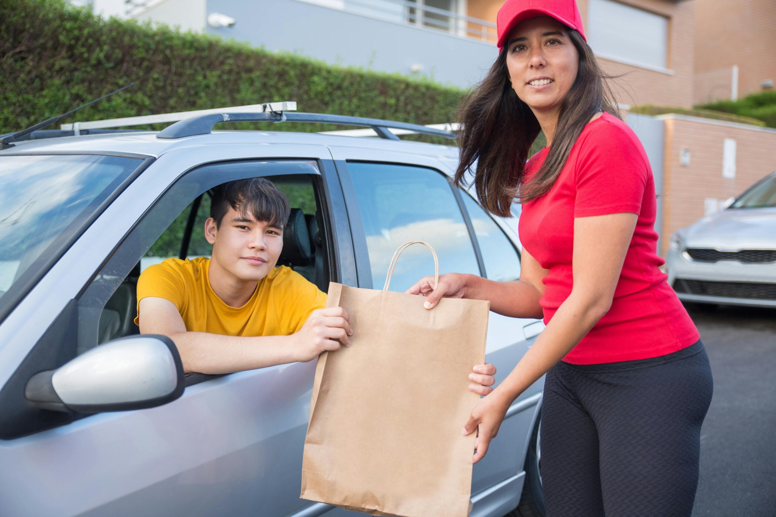 A woman in a red cap and shirt hands a brown paper bag to a young man sitting in a car. The man is reaching out through the window to receive the bag. They are outdoors in a residential area.
