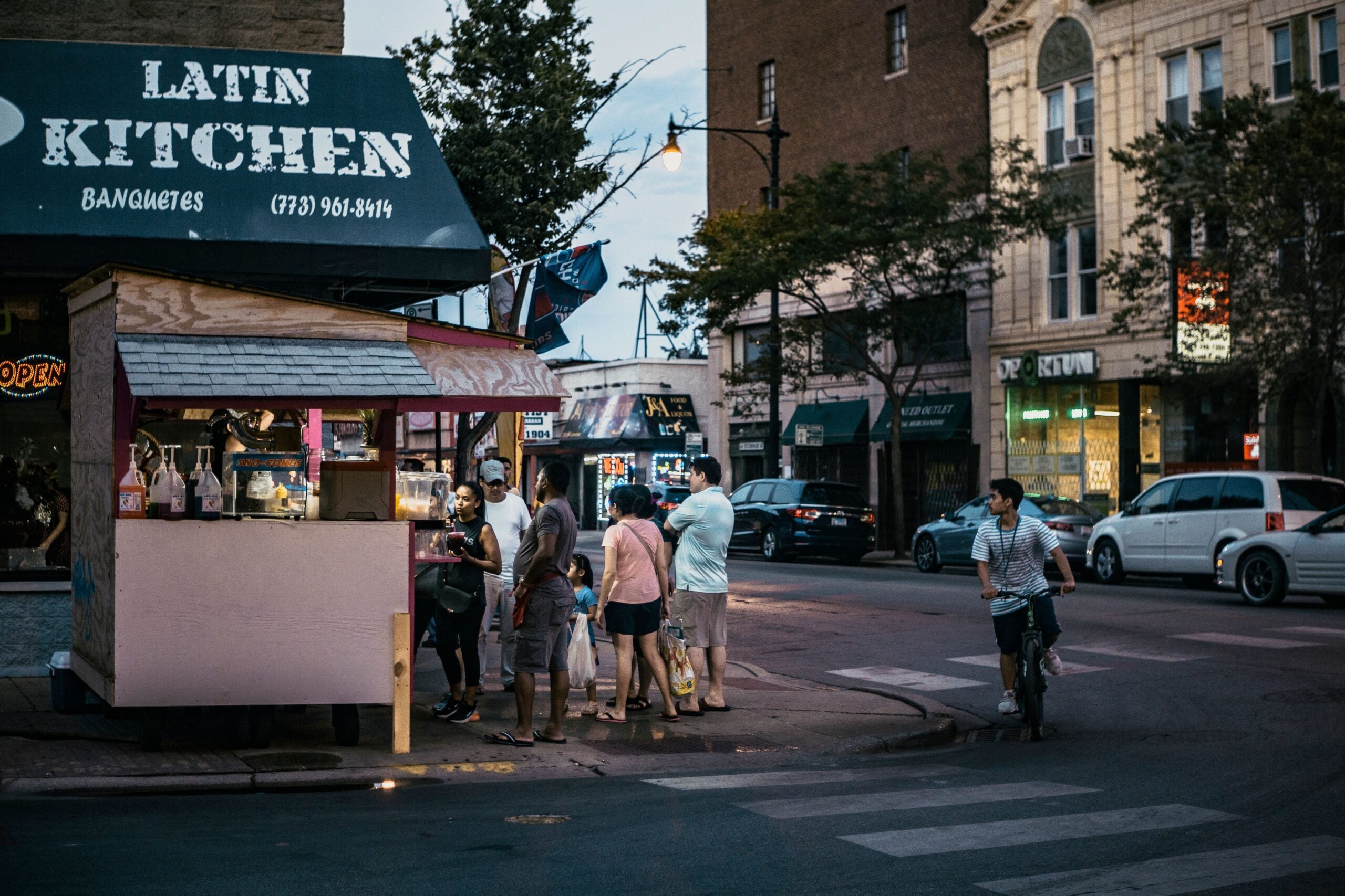 A group of people waits at a small food stand labeled "Latin Kitchen" on a city street corner at dusk, with surrounding buildings and a cyclist nearby.