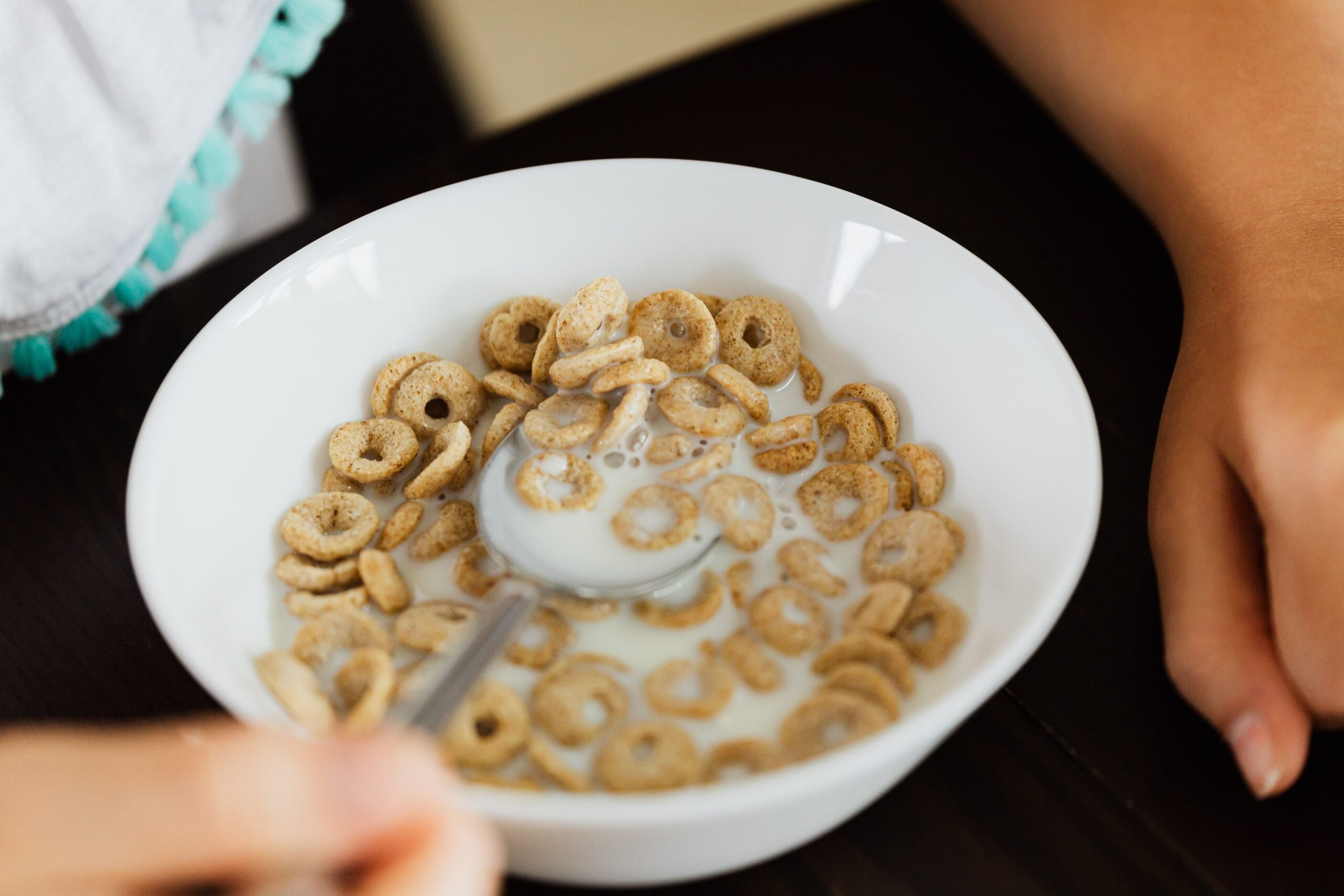 A close-up of a white bowl filled with ring-shaped cereal and milk, with a person holding a spoon and another hand resting on the dark table beside the bowl.