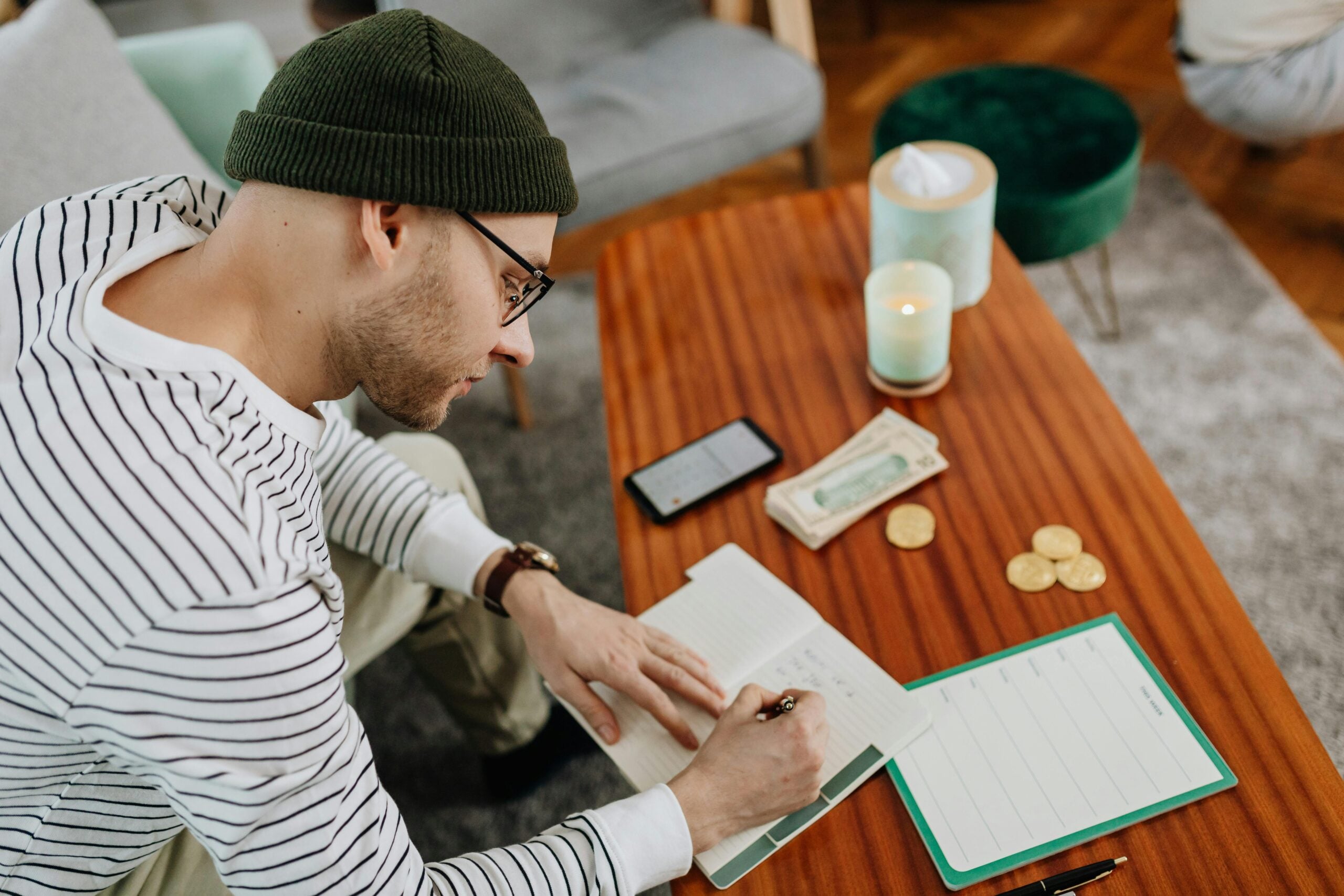 A person in a striped shirt and beanie writes in a notebook at a wooden table with cash, coins, a checklist, a phone, and lit candles. The setting appears to be a cozy living room.