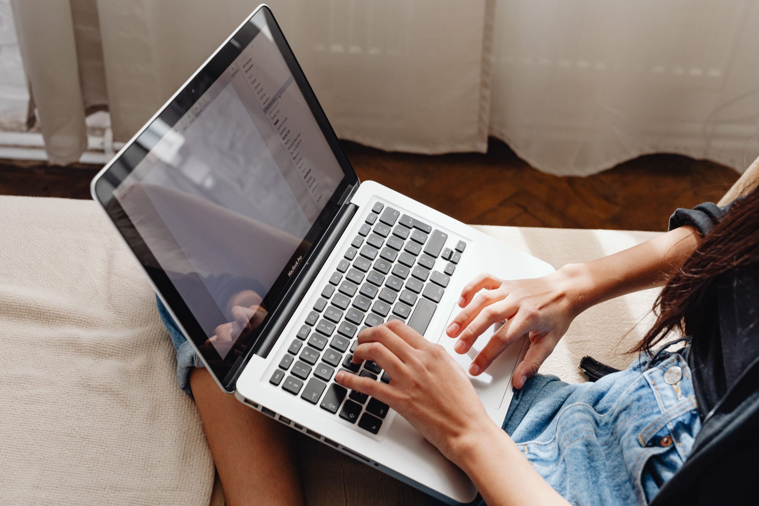A person sitting on a sofa types on a laptop resting on their lap. They are wearing a denim skirt and a dark top. The screen displays a document with text. Curtains and a wooden floor are visible in the background.