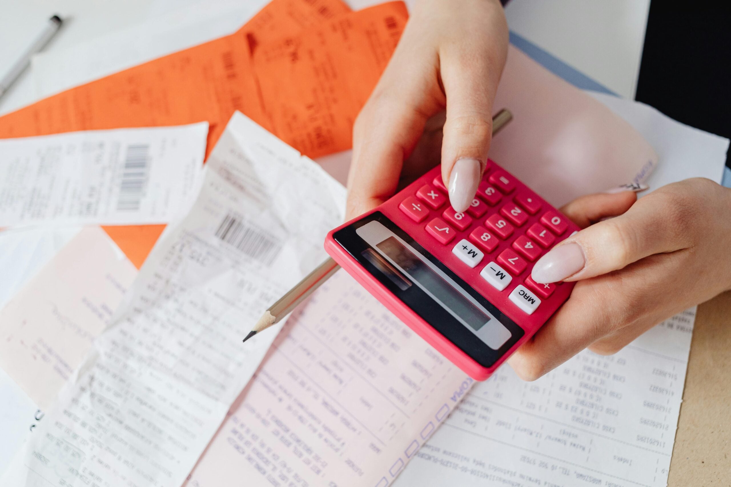 A person holding a pink calculator and pressing buttons, surrounded by several receipts, documents, and a pen on a table.