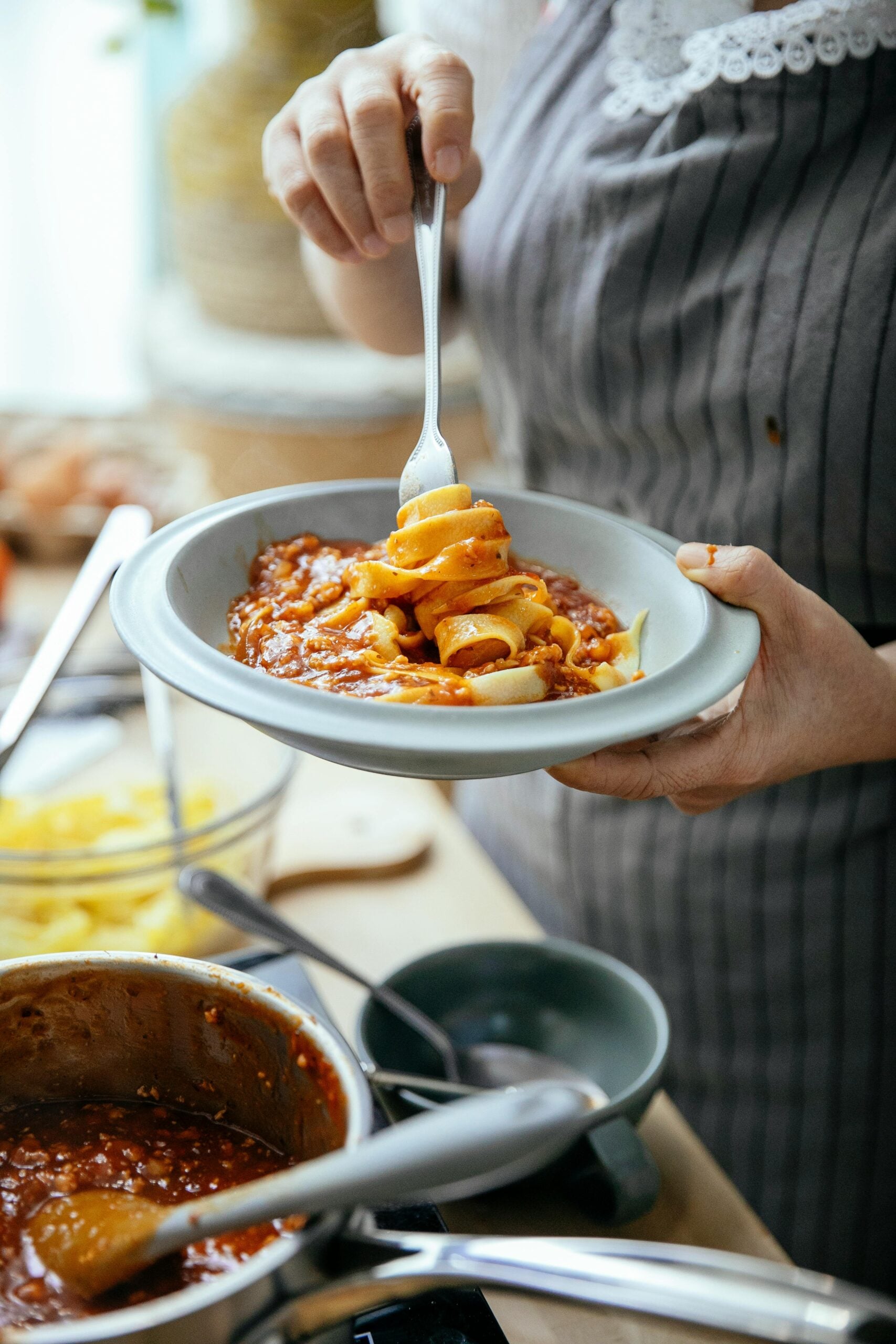 A person in a striped apron is holding a bowl of pasta with tomato sauce, twirling the pasta with a fork. Cookware and a pot of sauce are visible on the countertop in the foreground.