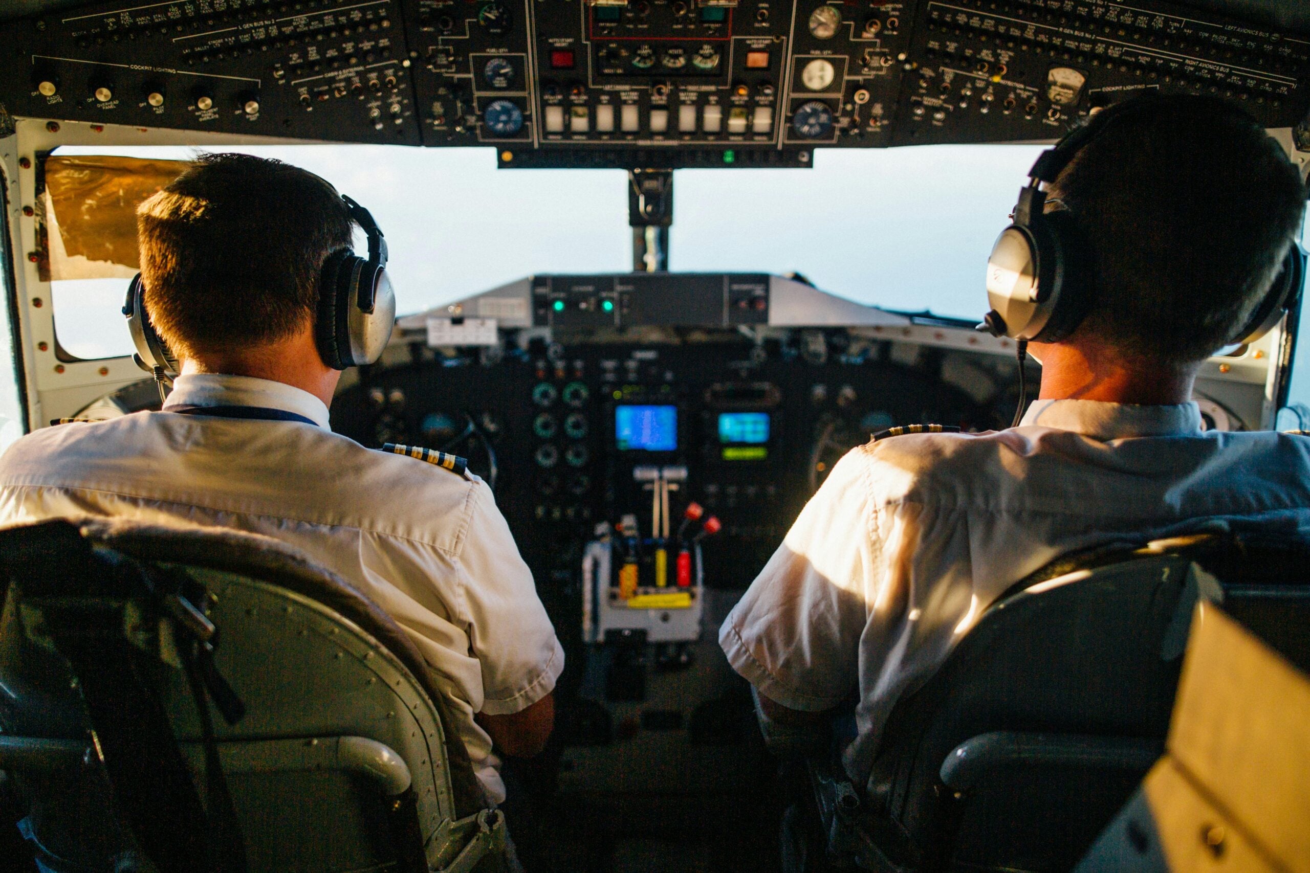 Two pilots wearing headsets sit in the cockpit of an airplane, facing the control panel and large windows, with various instruments and controls visible in front of them.