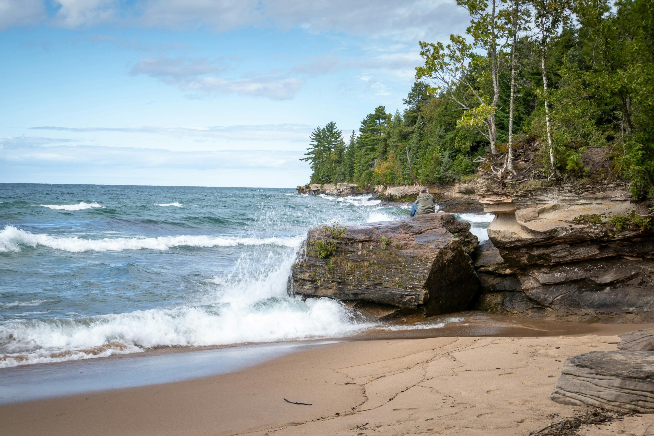Waves crash against rocky cliffs and sandy shore, with a person sitting on a large rock. Green trees line the coast under a partly cloudy sky, creating a peaceful lakeside scene.