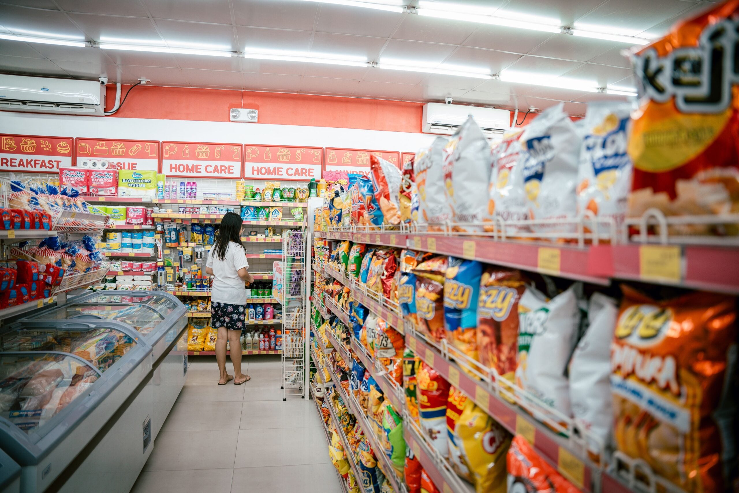A woman stands in an aisle of a brightly lit convenience store, looking at shelves stocked with various products. Snack bags and frozen items line the foreground and sides.