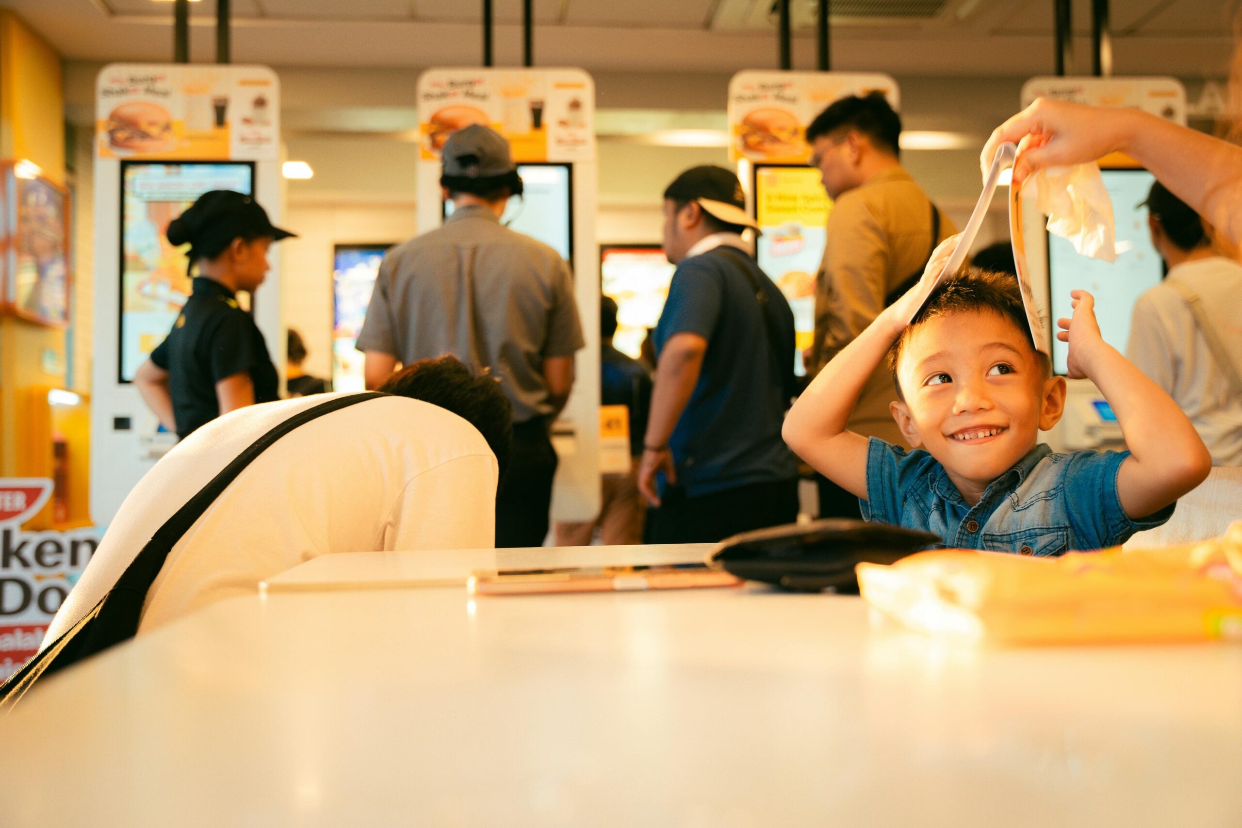 A young boy smiles playfully while holding a food wrapper above his head at a fast food restaurant, with several people standing in line and ordering at the counter in the background.