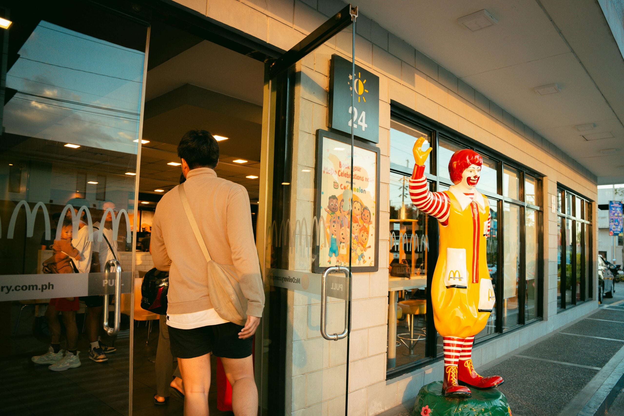 A person in a beige jacket and shorts enters a McDonald’s restaurant as a Ronald McDonald statue waves near the entrance on a sunny day.