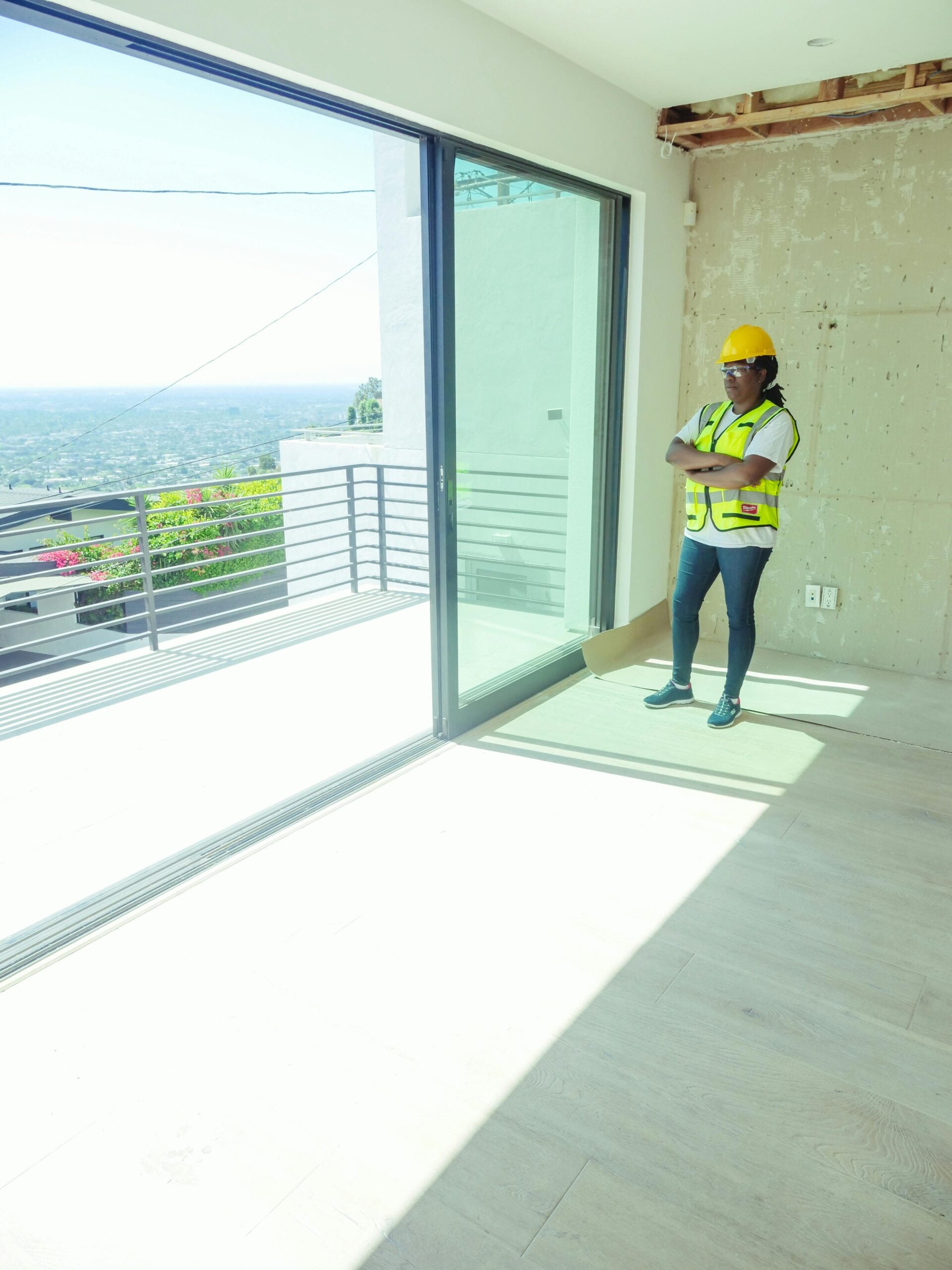 A construction worker in a yellow hard hat and safety vest stands with arms crossed inside a bright, unfinished room with large sliding glass doors—a setting that highlights the promise of Cost of Living Relief through new, affordable housing.