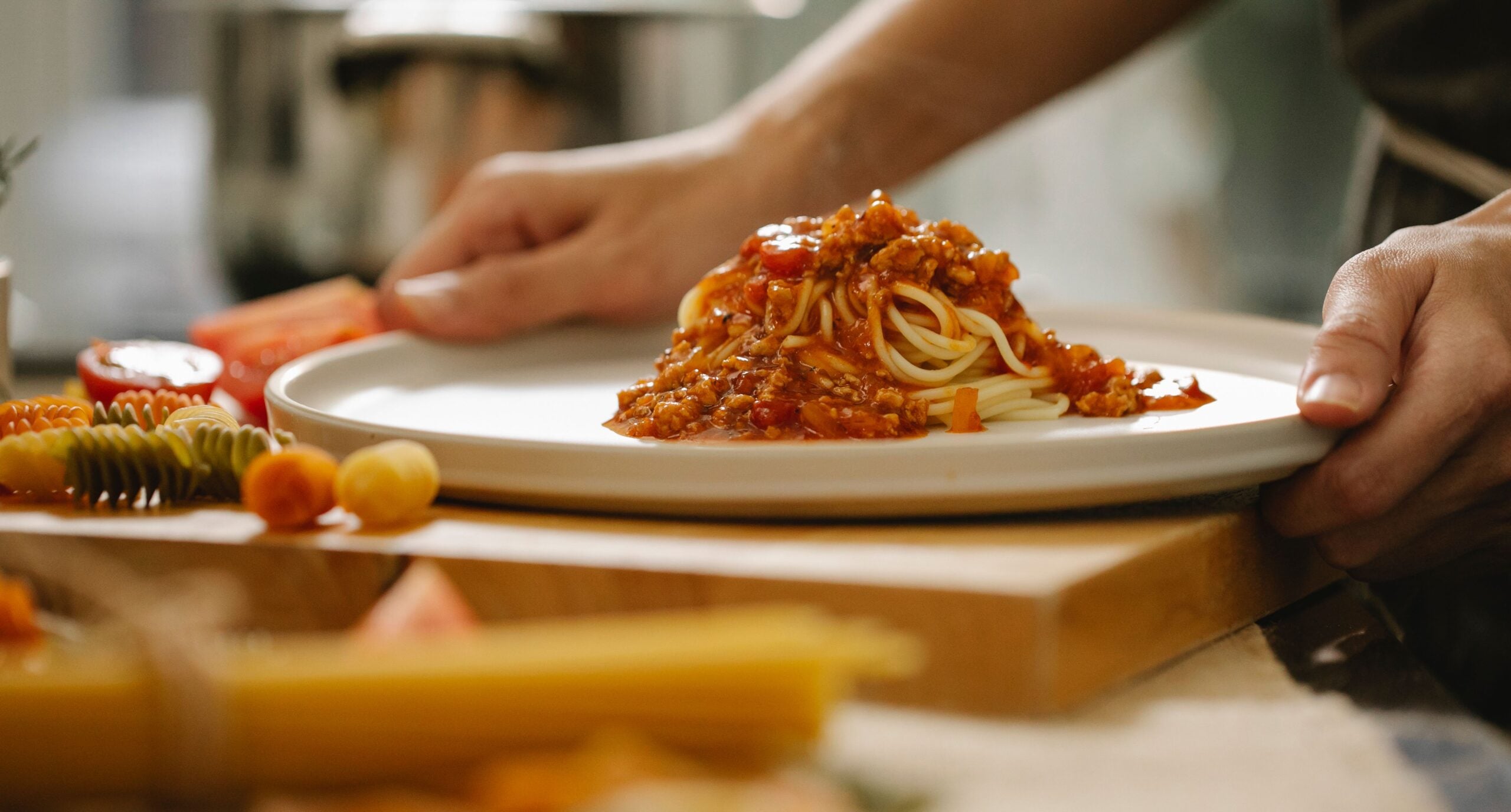 A person placing a white plate with spaghetti topped with tomato meat sauce on a wooden surface, surrounded by uncooked pasta and vegetables.