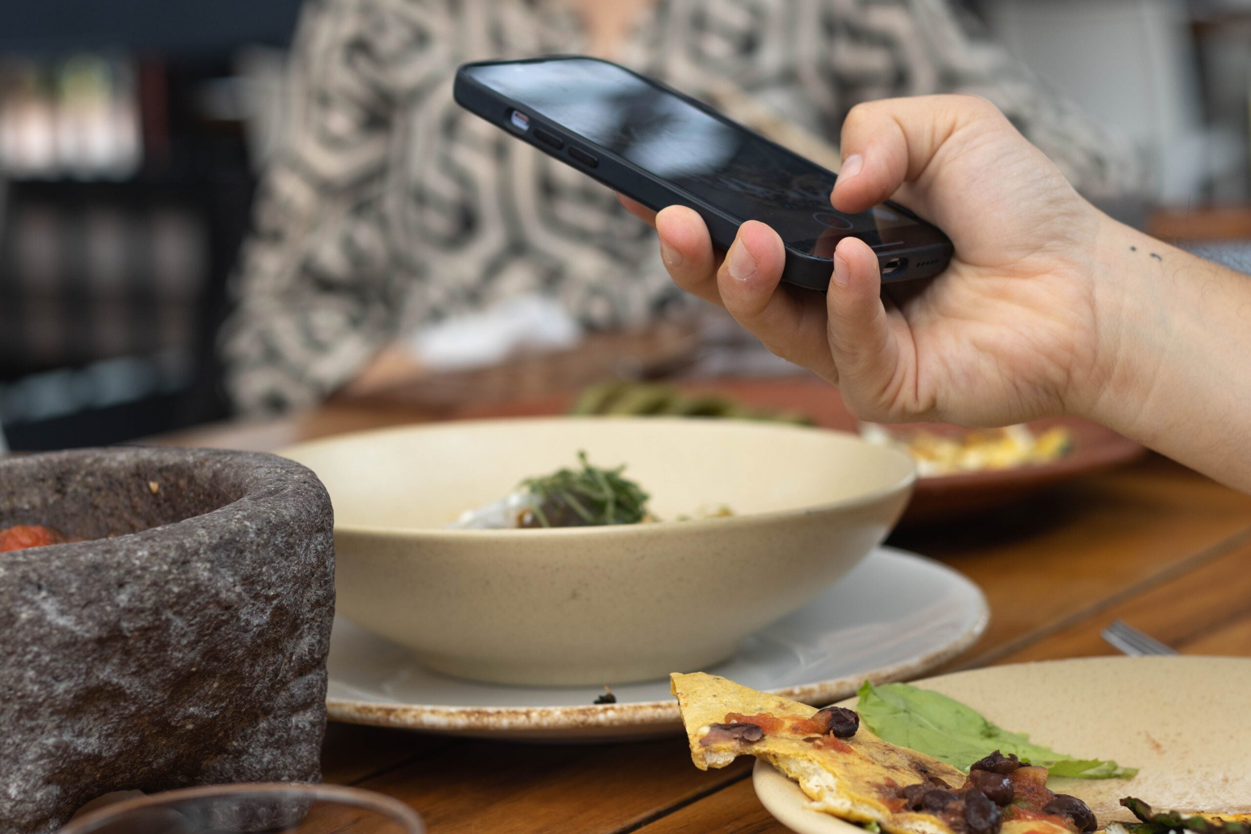 A person holds a smartphone above a dining table with bowls and plates of food, including a slice of bread with beans. The focus is on the hand and phone, with other diners and dishes slightly blurred in the background.