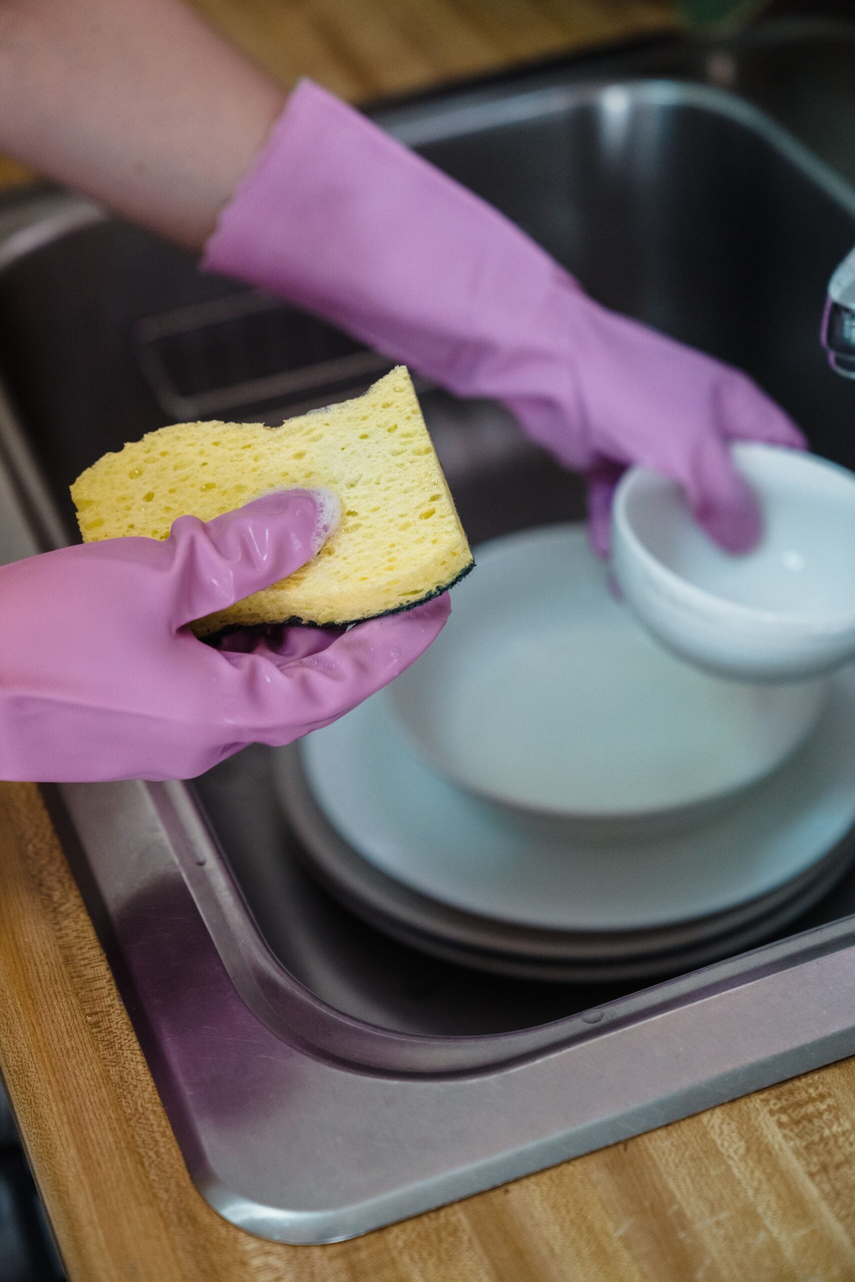 A person wearing pink rubber gloves holds a yellow sponge and a white bowl over a sink filled with dishes, preparing to wash them.