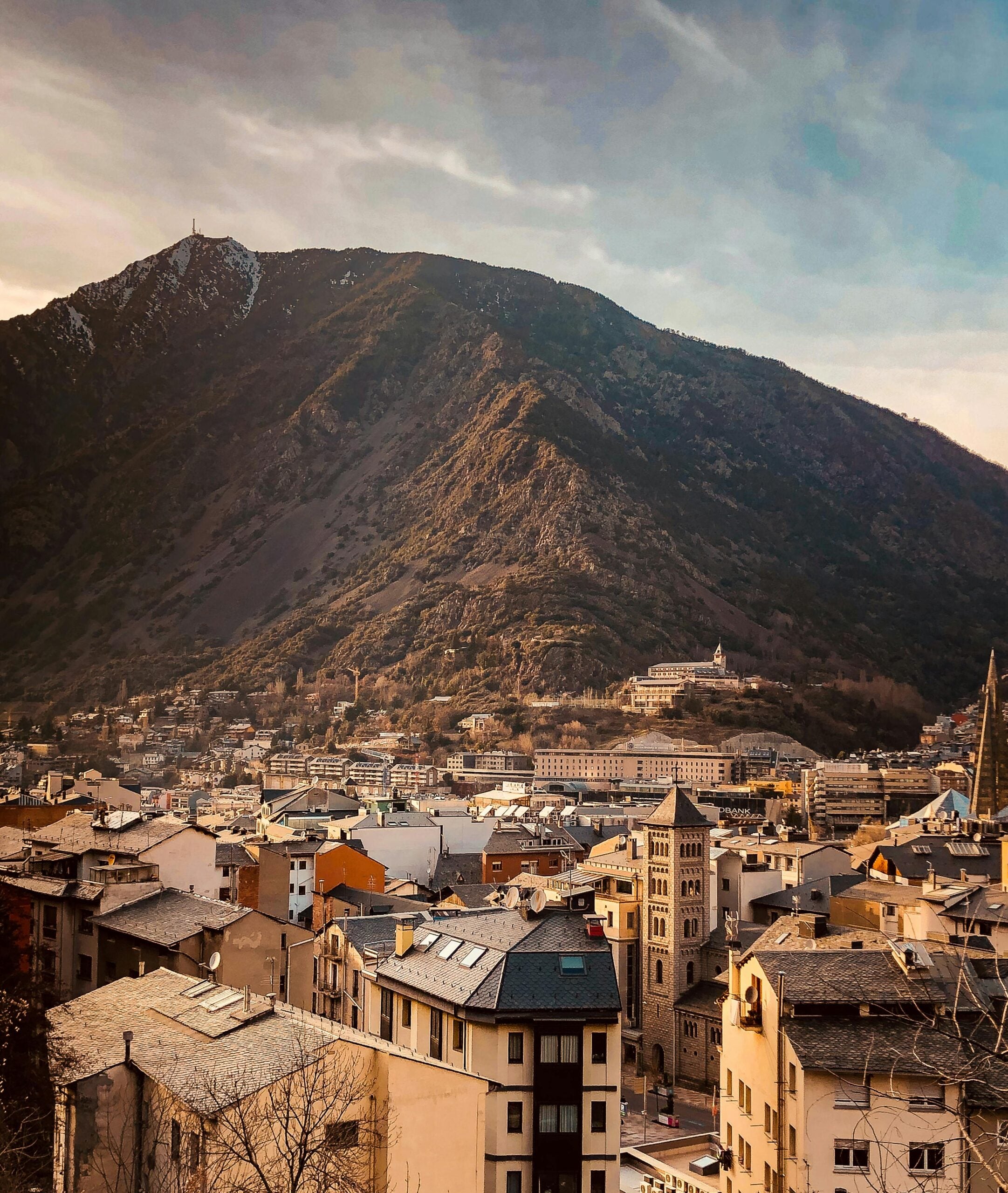 A picturesque view of a town with stone buildings and rooftops, set against a large, tree-covered mountain under a blue sky with wispy clouds at sunset or sunrise.