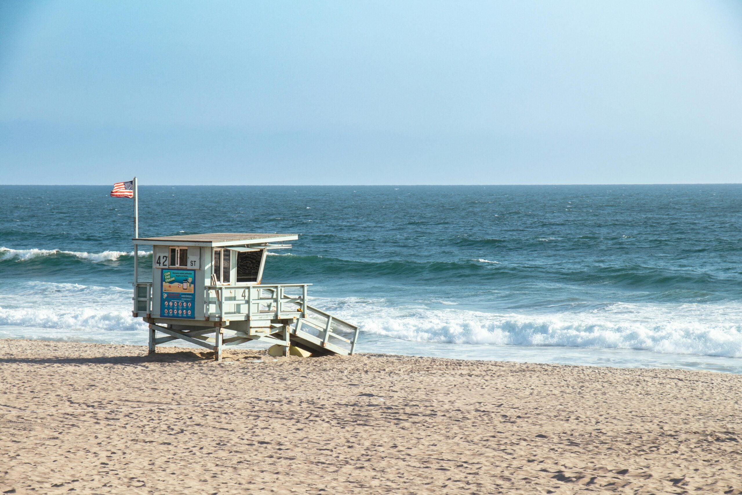 A lifeguard tower with an American flag stands on a sandy beach overlooking blue ocean waves under a clear sky.