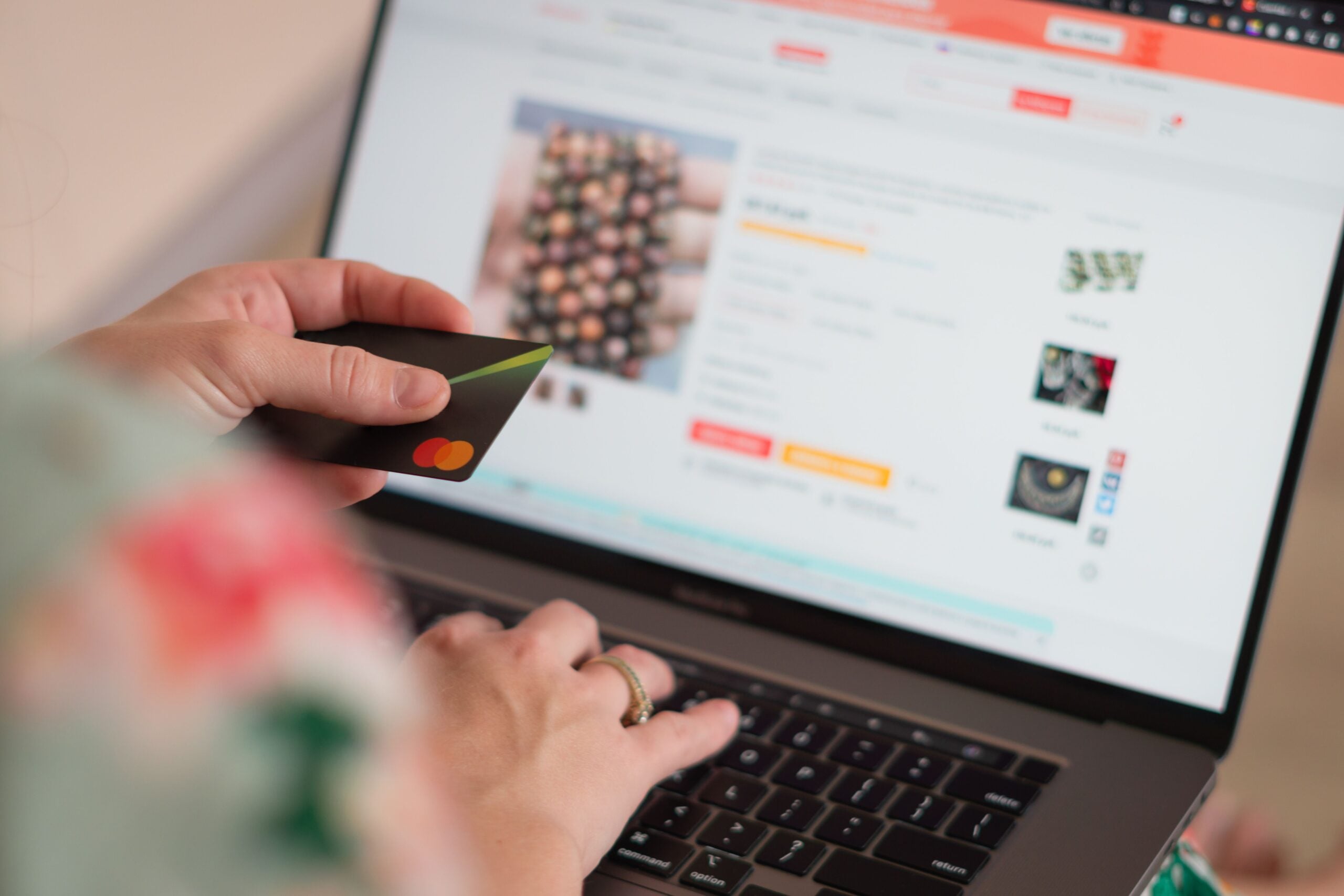 A person holding a credit card in one hand and typing on a laptop with the other, shopping for jewelry online as product images and prices are visible on the screen.