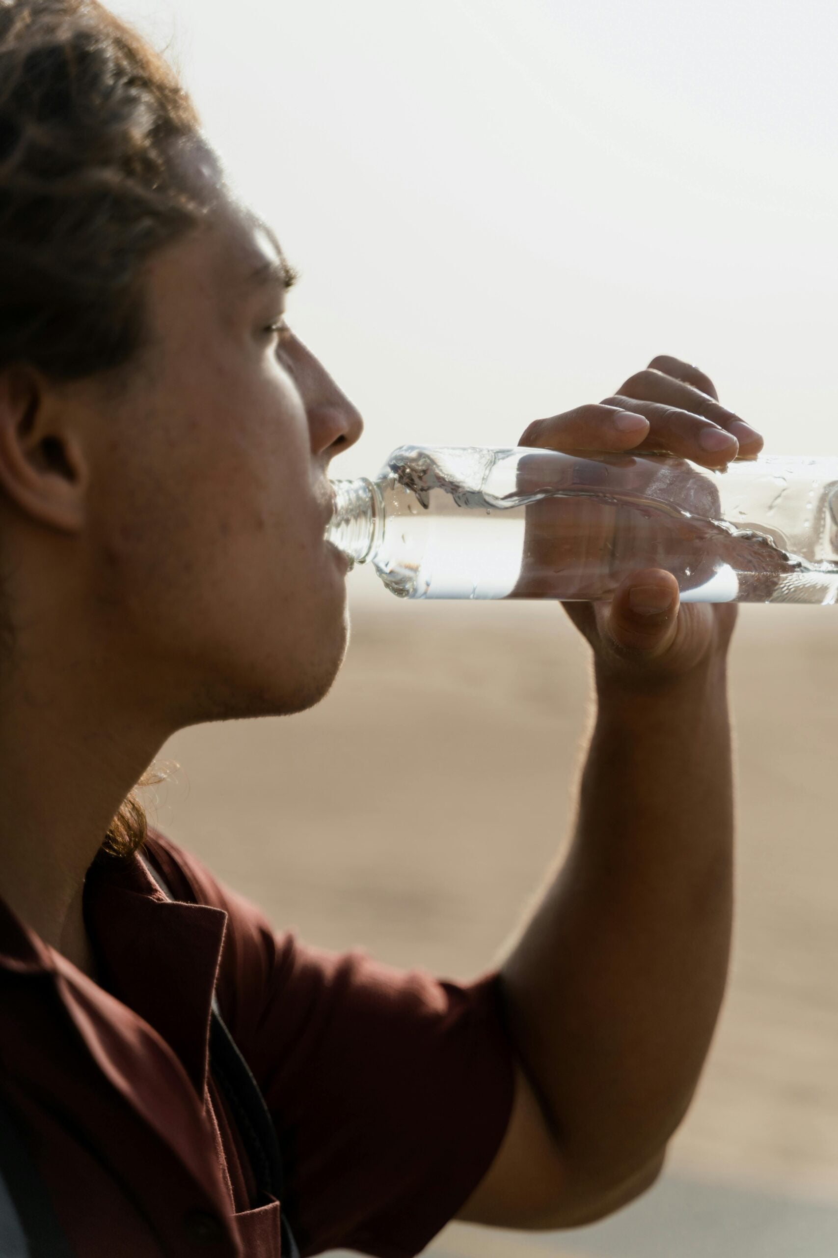 A person with long hair drinks water from a clear plastic bottle outdoors, with a blurred sandy or desert-like background visible.