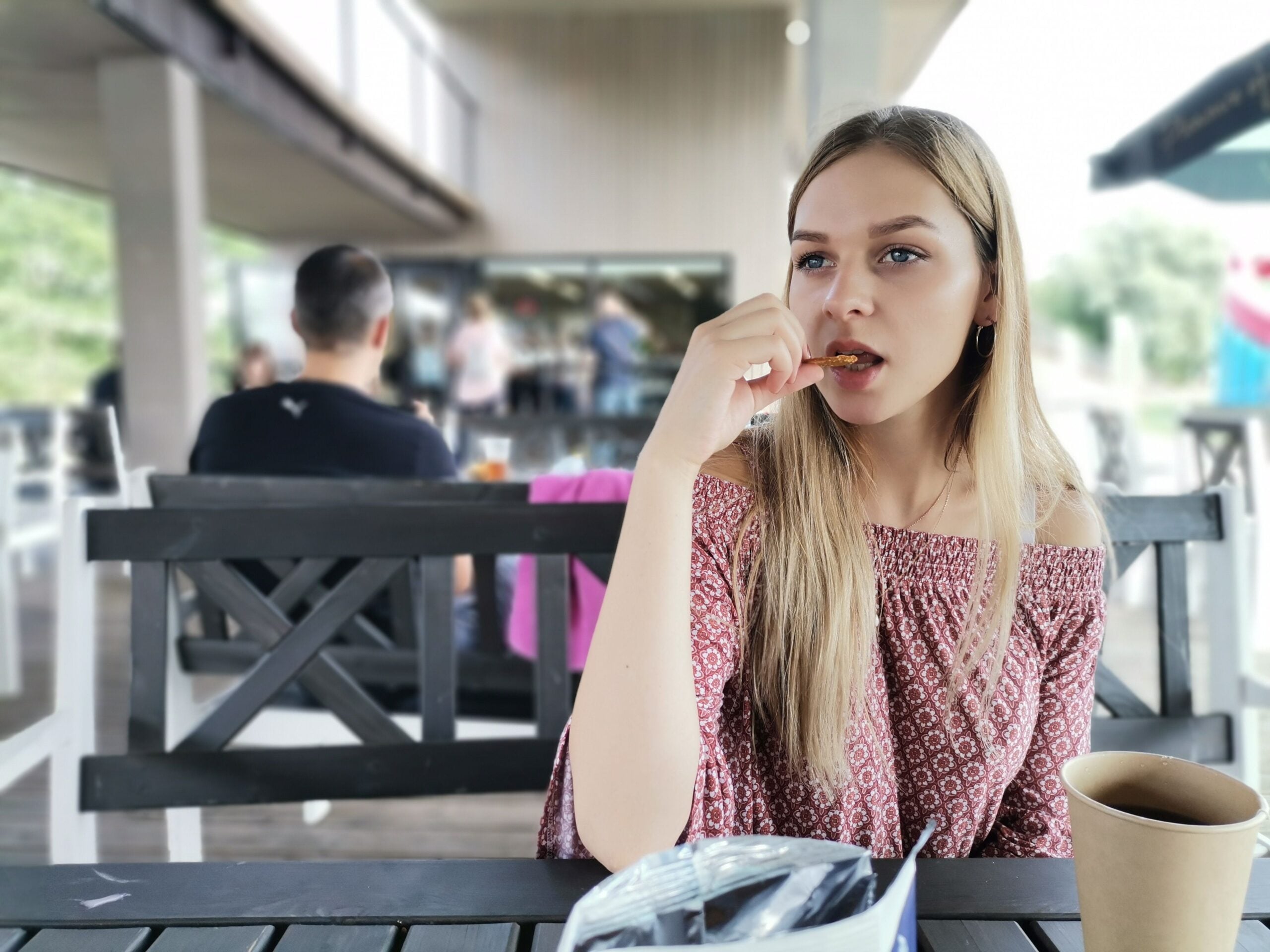 A young woman with long blonde hair sits at an outdoor café table, eating a snack. She is wearing an off-the-shoulder patterned top. A paper cup and food wrapper are on the table. The background shows other people and a building.