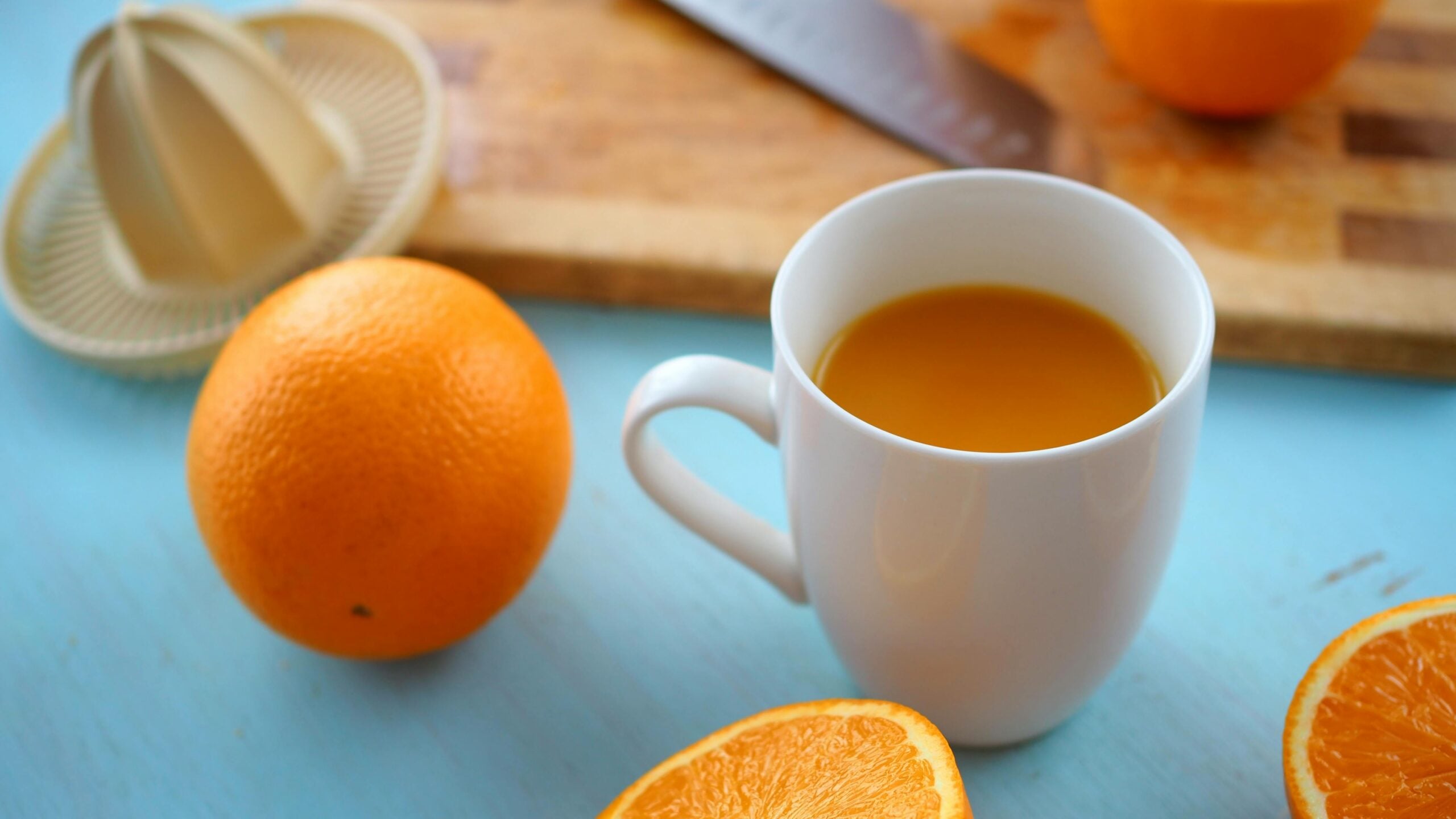 A white mug filled with orange juice sits on a blue surface next to a whole orange, a sliced orange, a manual juicer, and a cutting board with a knife.