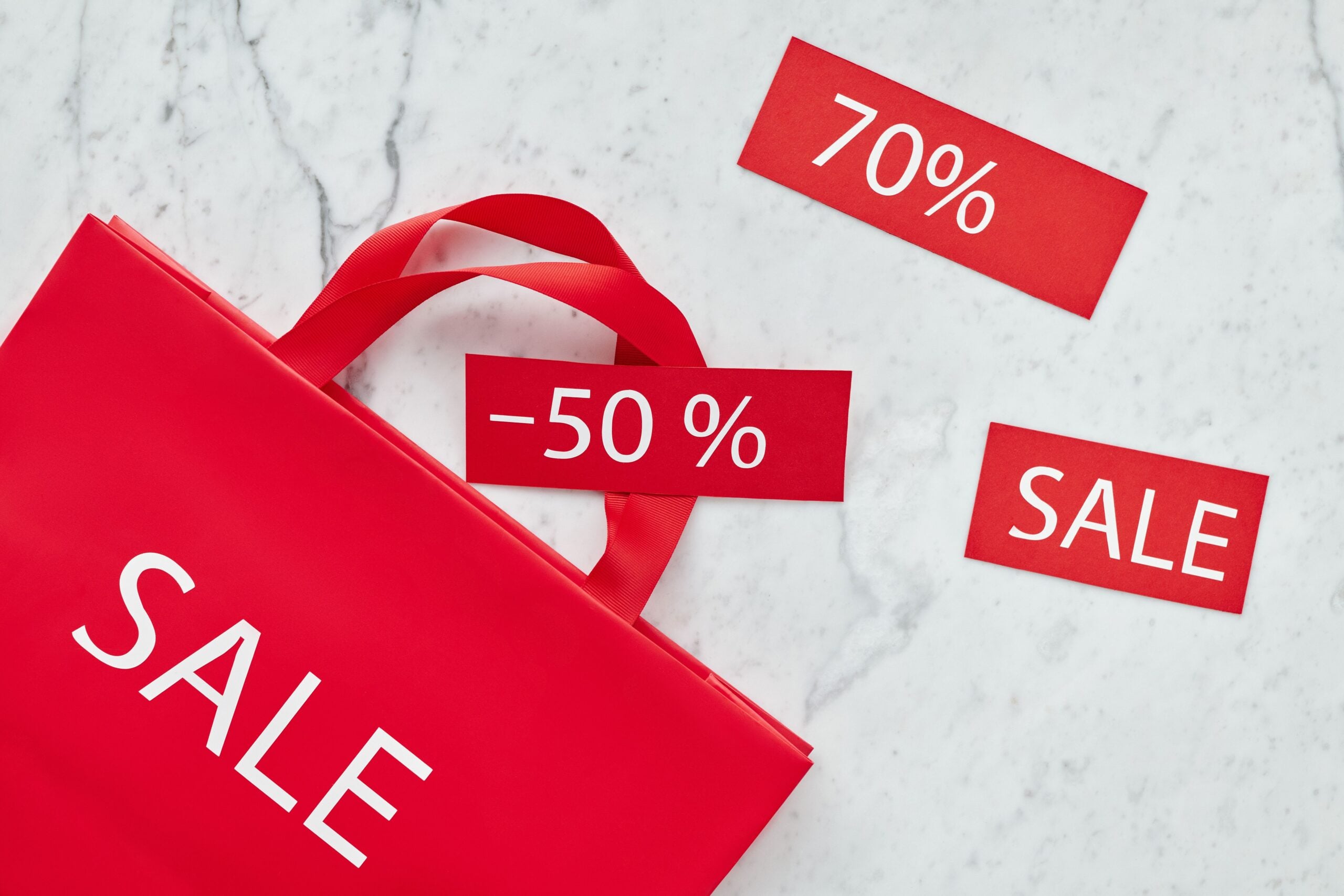 A red shopping bag labeled "SALE" sits on a marble surface, surrounded by red cards with white text showing "70%", "-50%", and "SALE," indicating discounts.
