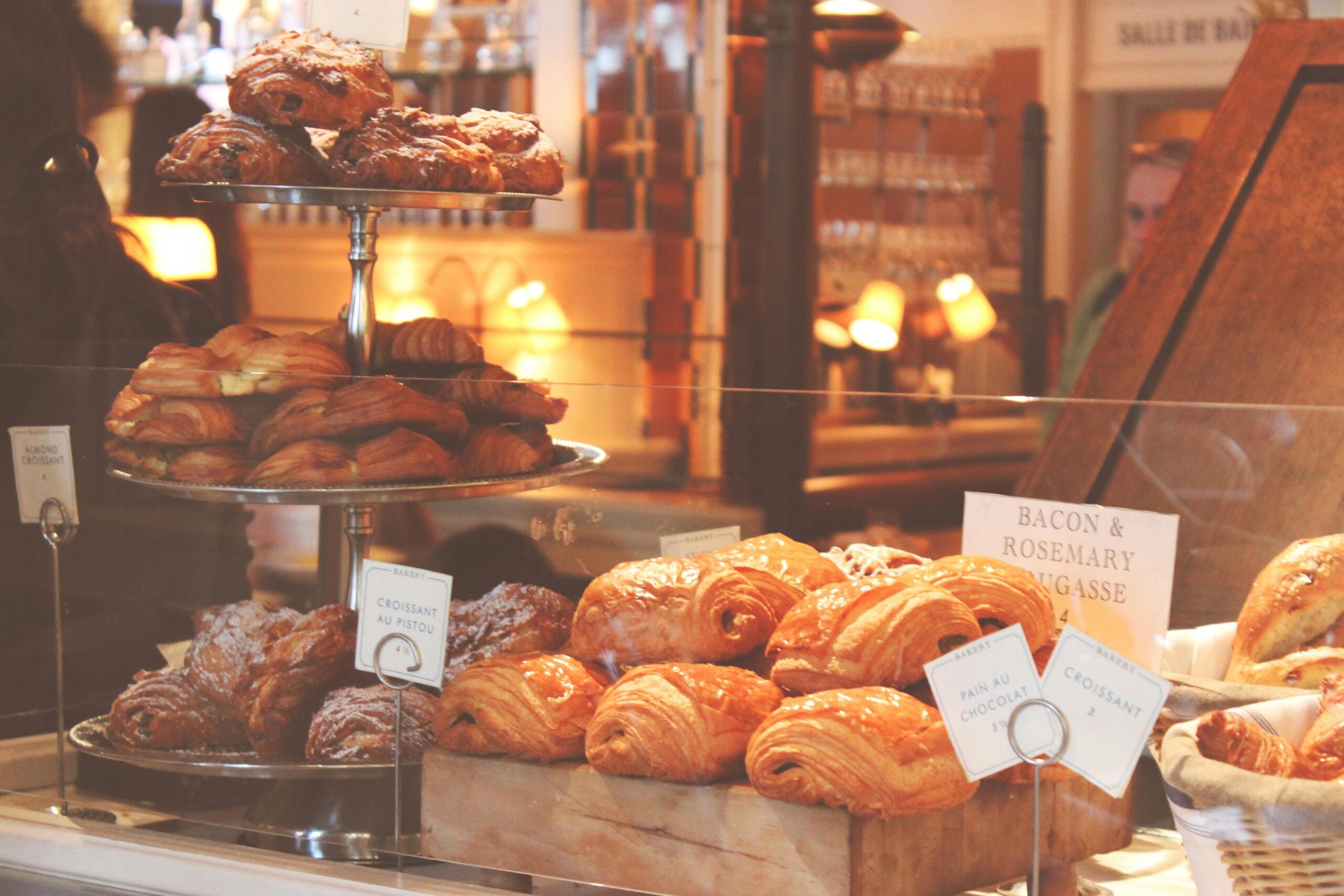 A bakery display filled with a variety of pastries, including croissants and pain au chocolat, behind a glass counter with labels. The warm lighting creates a cozy atmosphere.