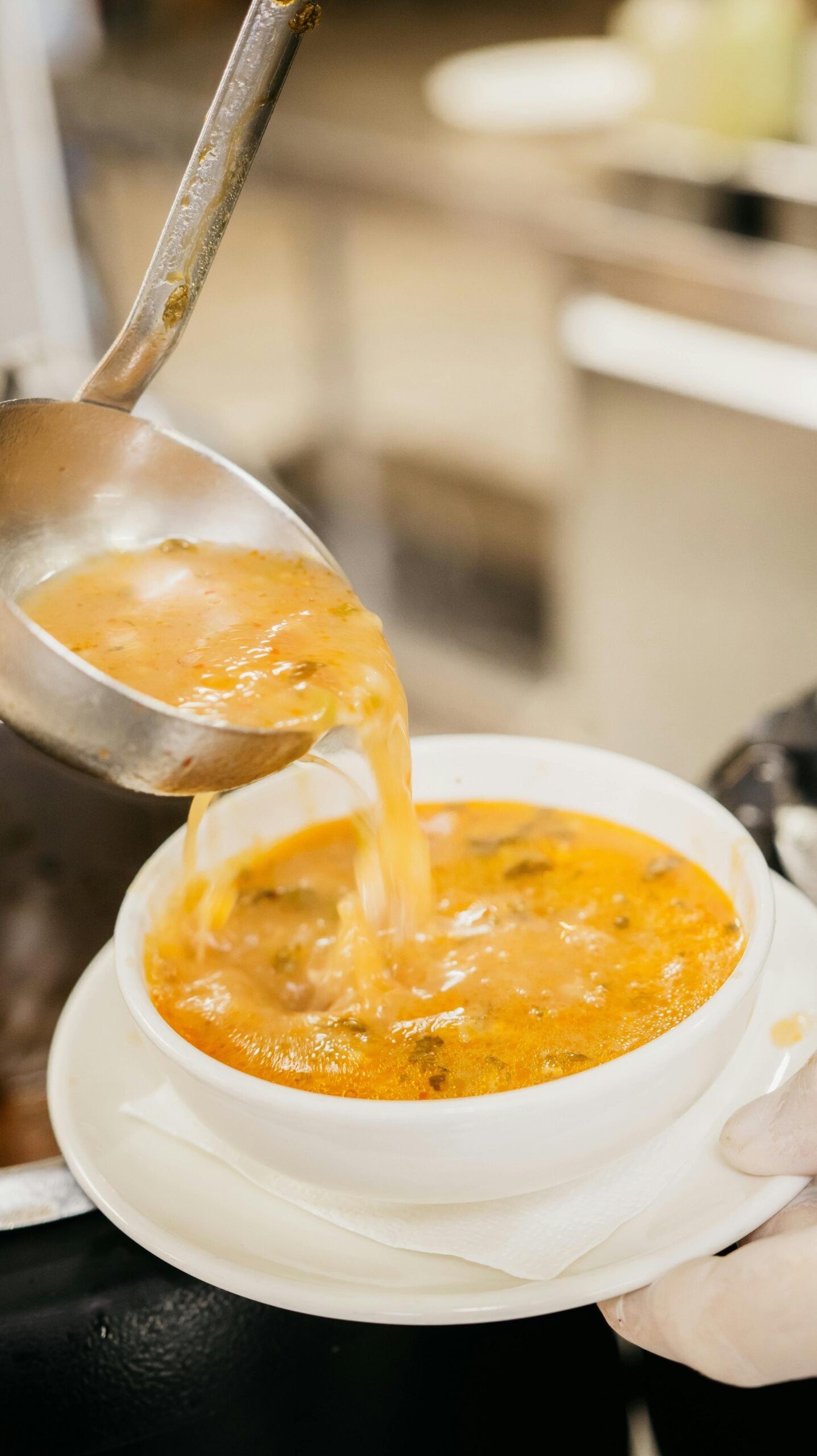 A close-up of a ladle pouring hot, orange-colored soup with herbs into a white bowl, which is placed on a white saucer. The setting appears to be a kitchen or a cafeteria.