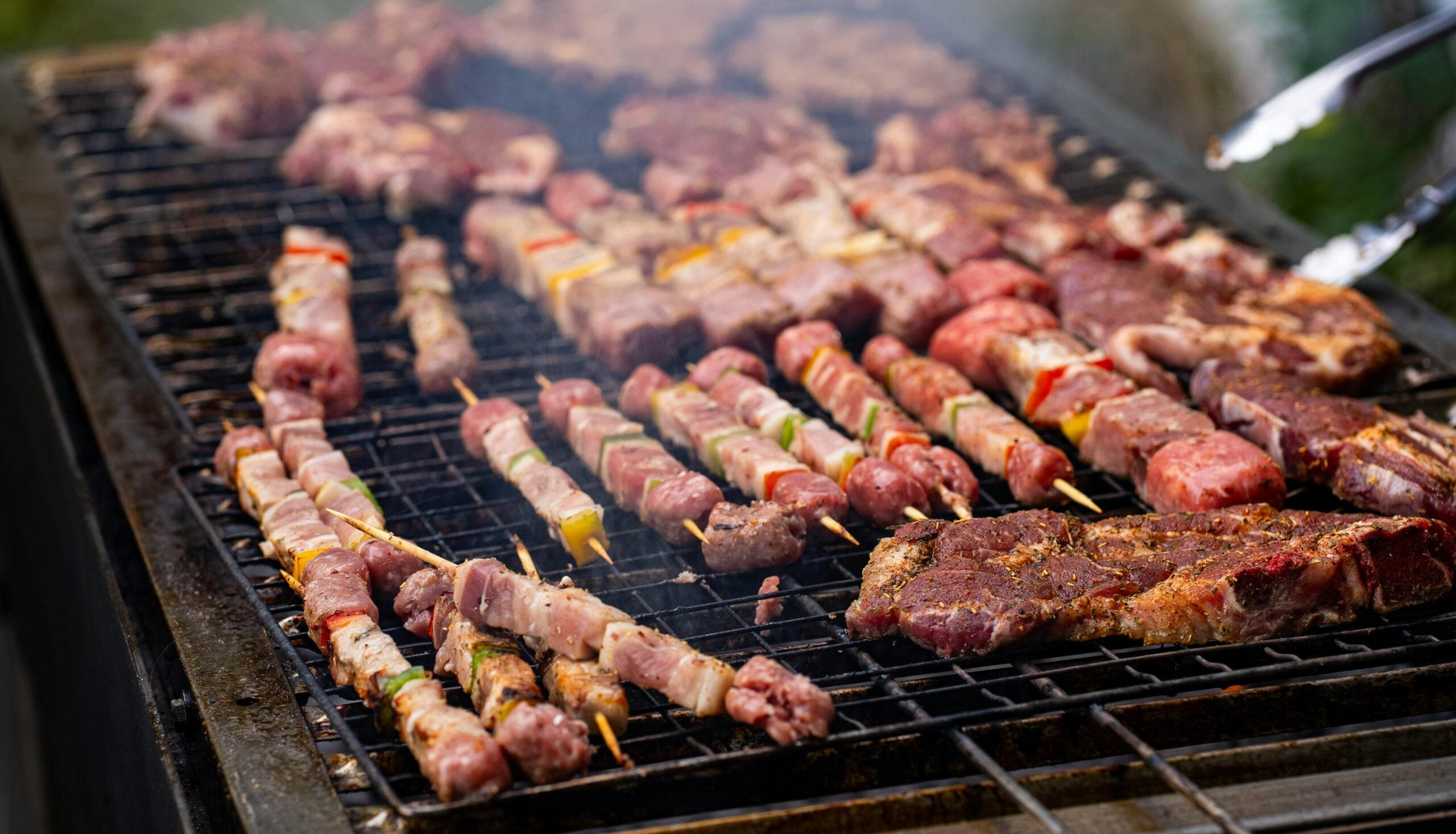 An assortment of meats and skewers with vegetables cooking on a smoky outdoor grill, with some pieces marinated and others unseasoned, all arranged closely together over the grates.