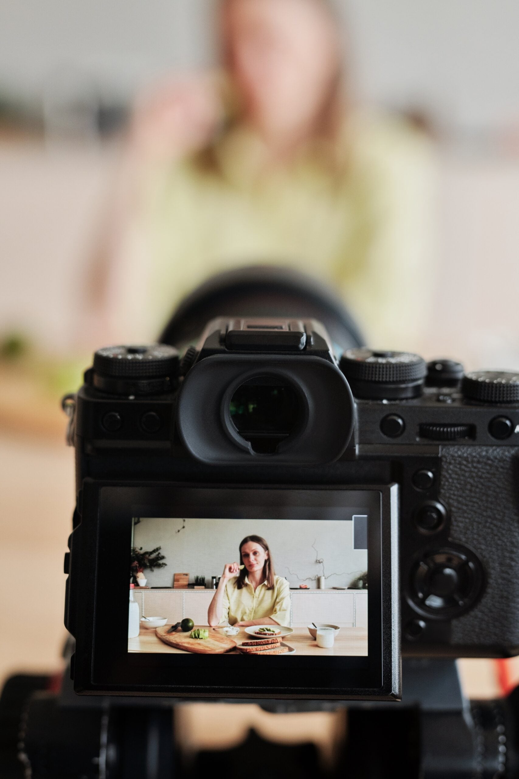 A digital camera captures a woman sitting at a table with food, as seen on the camera’s display screen. The woman is slightly out of focus in the background.