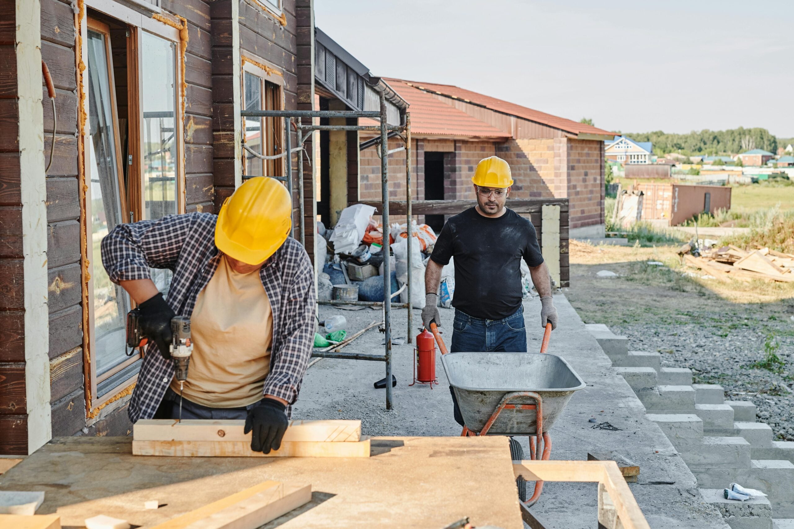 Two construction workers in yellow hard hats work outside a building; one uses a power drill on wood at a table, while the other stands nearby with a wheelbarrow, highlighting how Cost of Living Relief supports those in trades.