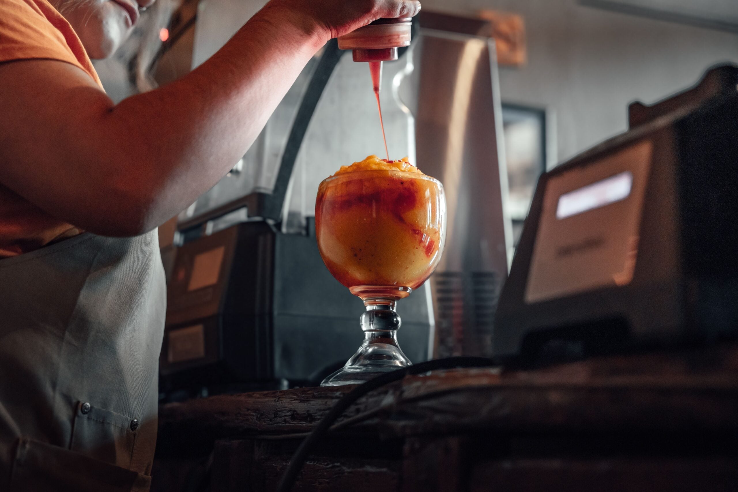 A person in an apron is squeezing syrup onto a large glass filled with a yellow and orange frozen dessert, possibly a slushie or sorbet, on a counter in a dimly lit setting.