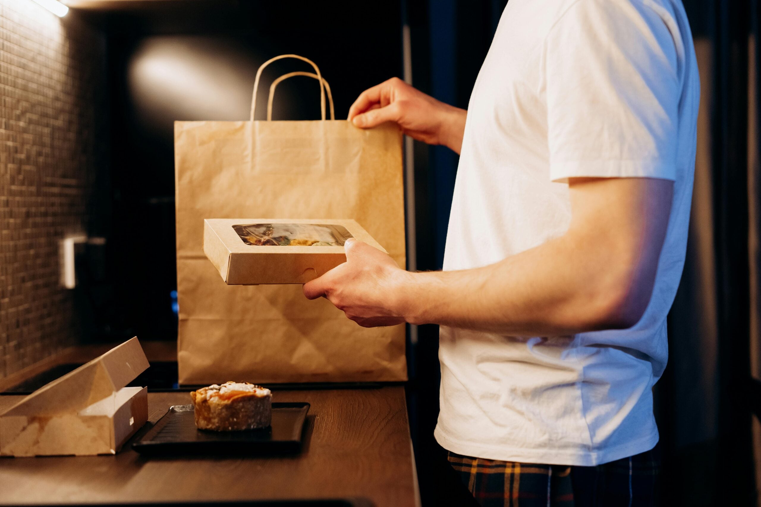 A person in a white t-shirt and plaid pants stands at a kitchen counter, taking food out of a paper bag and holding a takeout container, with more food and a tray on the counter.
