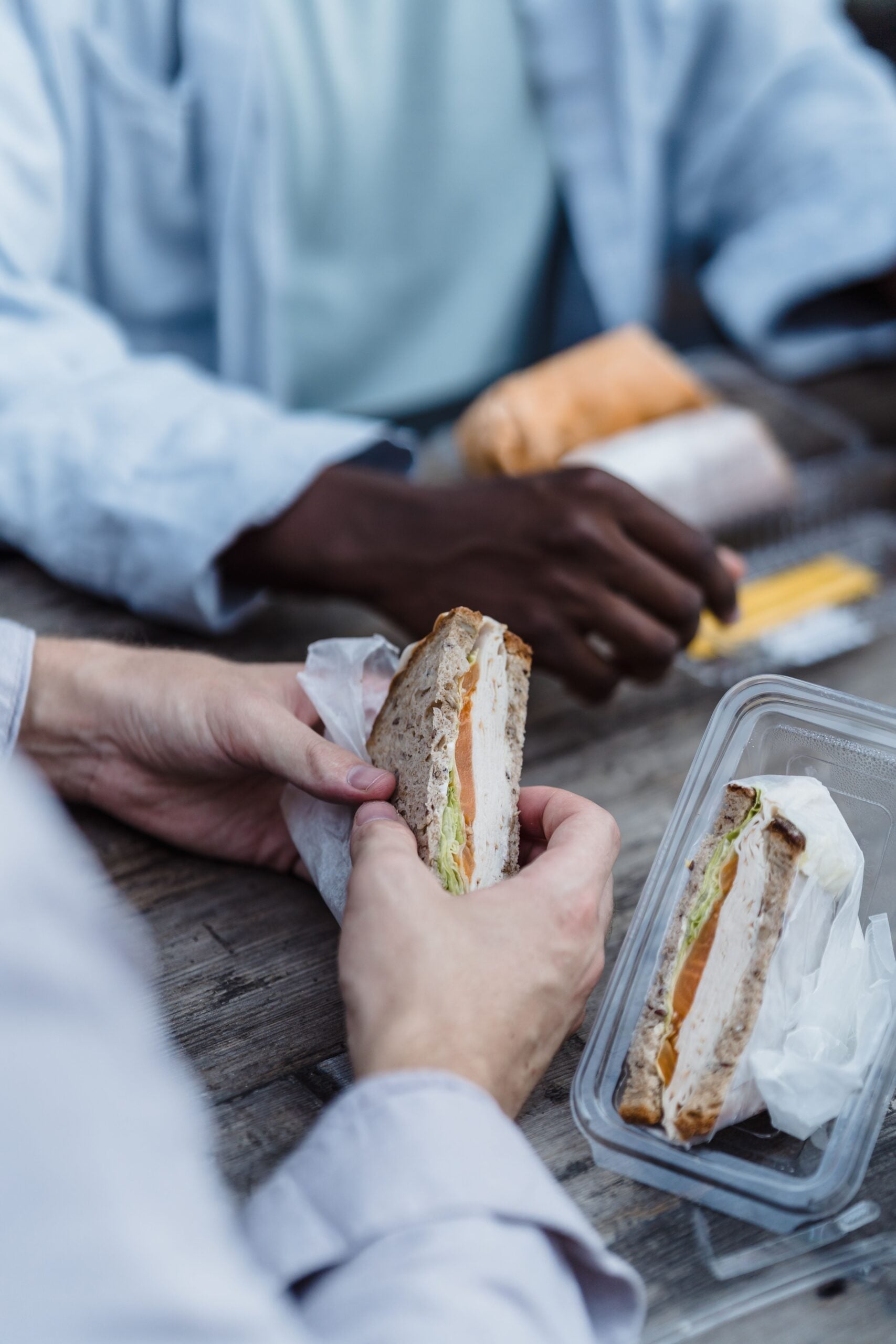 Two people sit at a wooden table holding sandwiches made with whole grain bread, lettuce, tomato, and cheese. One sandwich is partially wrapped in paper, and another sits in a clear plastic container nearby.