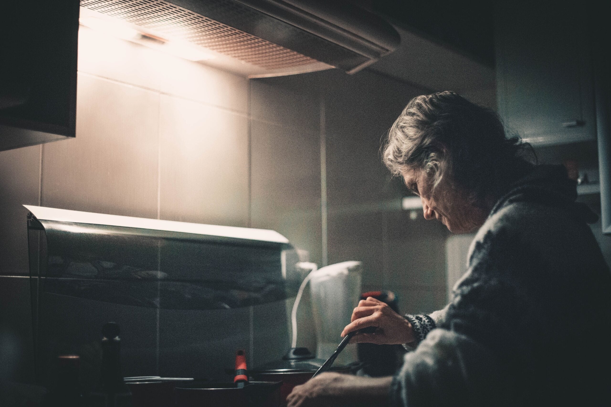 An older woman with gray hair is cooking at a stove in a dimly lit kitchen, concentrating as she stirs food in a pot. Warm light shines above her, creating a cozy atmosphere.