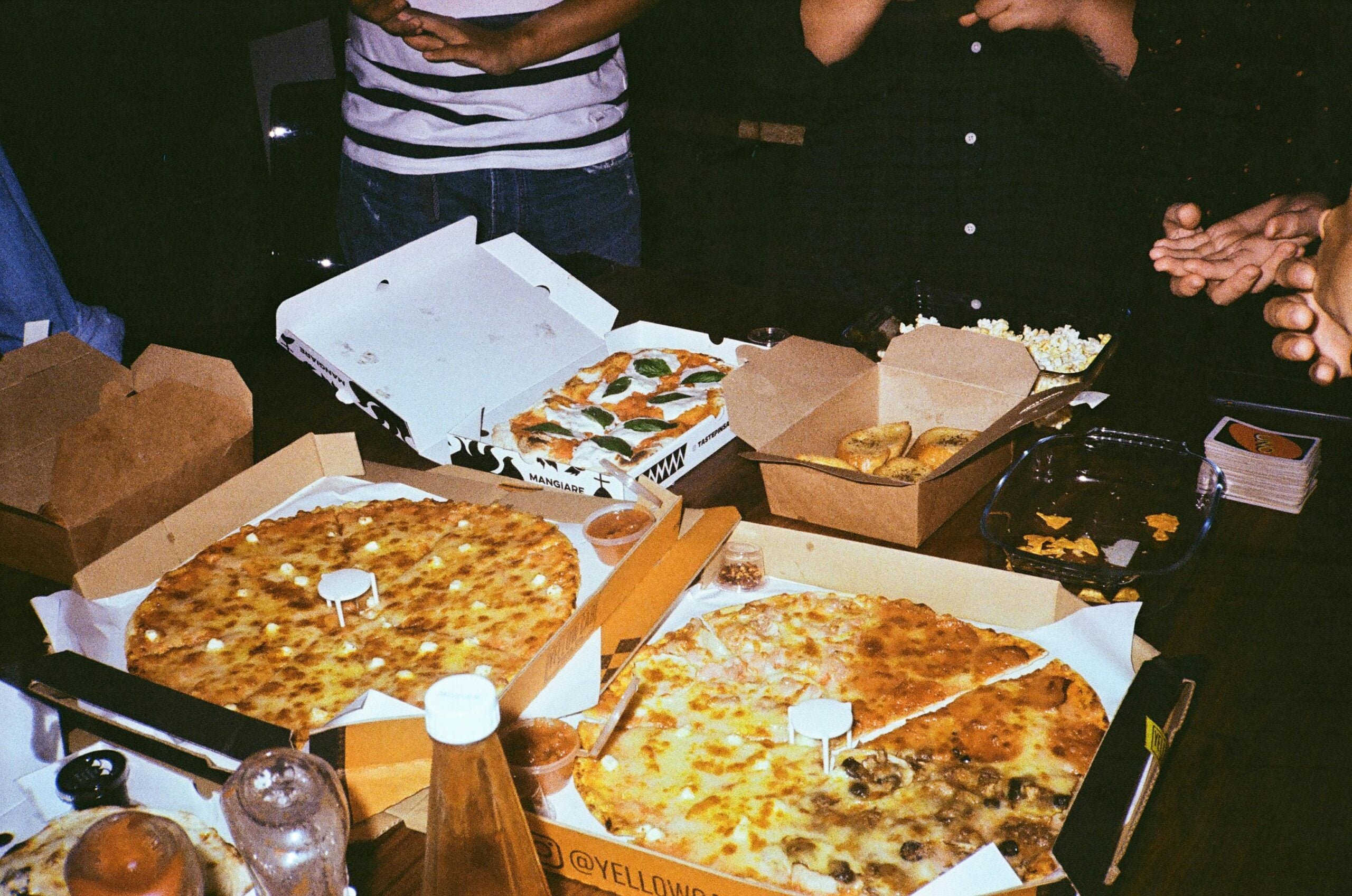 Several people stand around a table filled with open pizza boxes, breadsticks, snacks, and drinks, suggesting a casual gathering or party. Only the people's hands and torsos are visible.