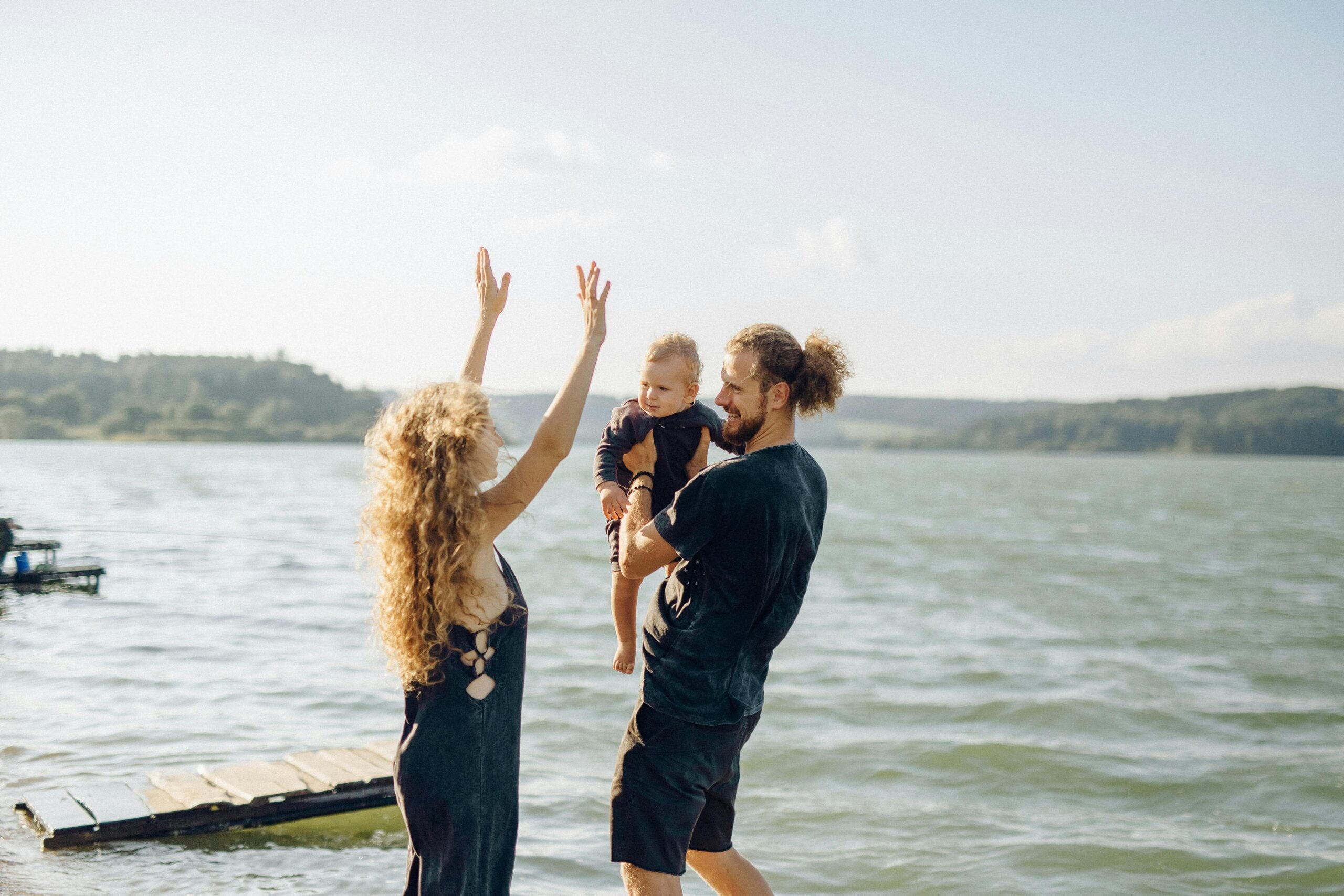 A woman with curly hair raises her arms joyfully beside a smiling man holding a toddler near a lake, with water and trees in the background on a sunny day.